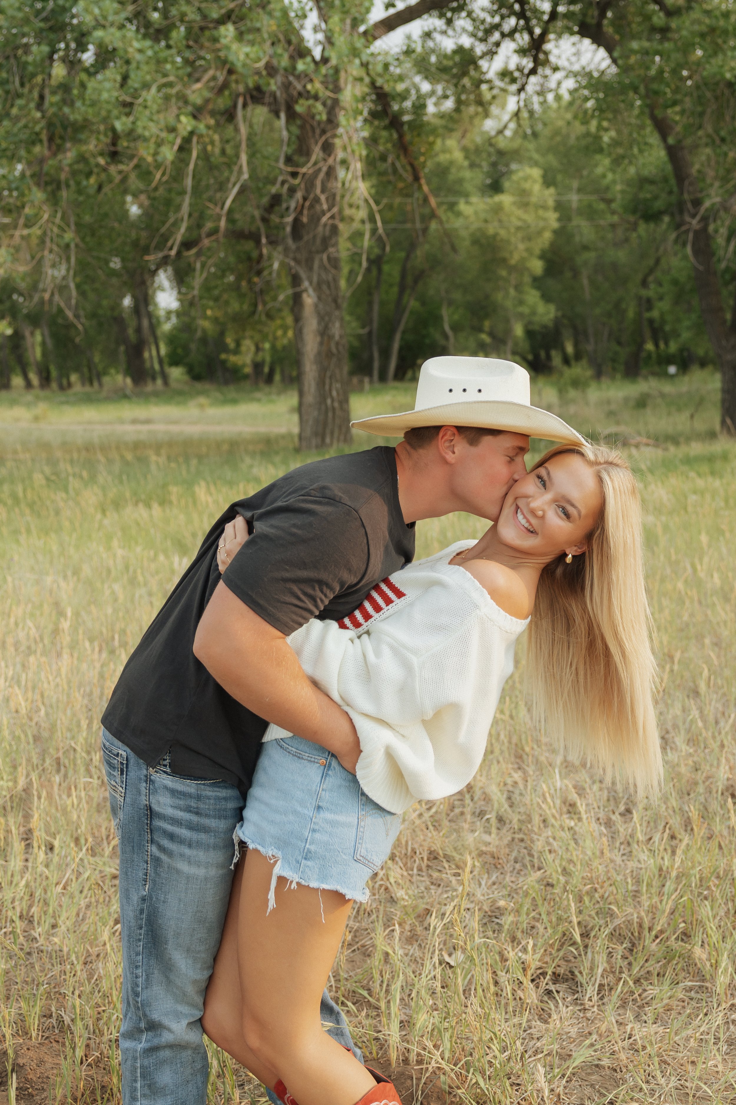 A young couple in a field surrounded by trees; the man is wearing a large white hat and dark shirt, and the woman is smiling and wearing a white sweater, denim shorts, and cowboy boots; the man is kissing the woman on the cheek.