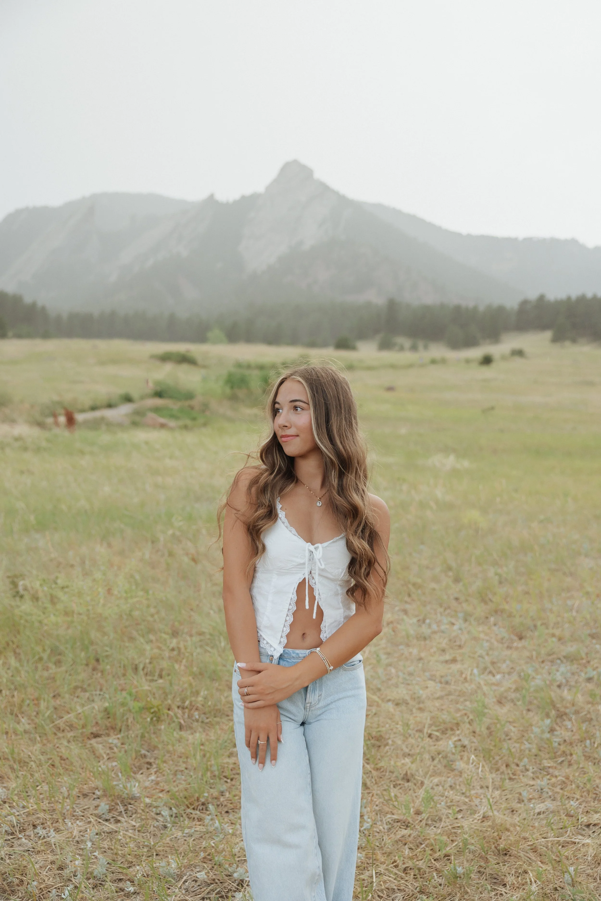 Young woman with long wavy hair wearing a white crop top and light blue jeans standing in a grassy field with mountains in the background.