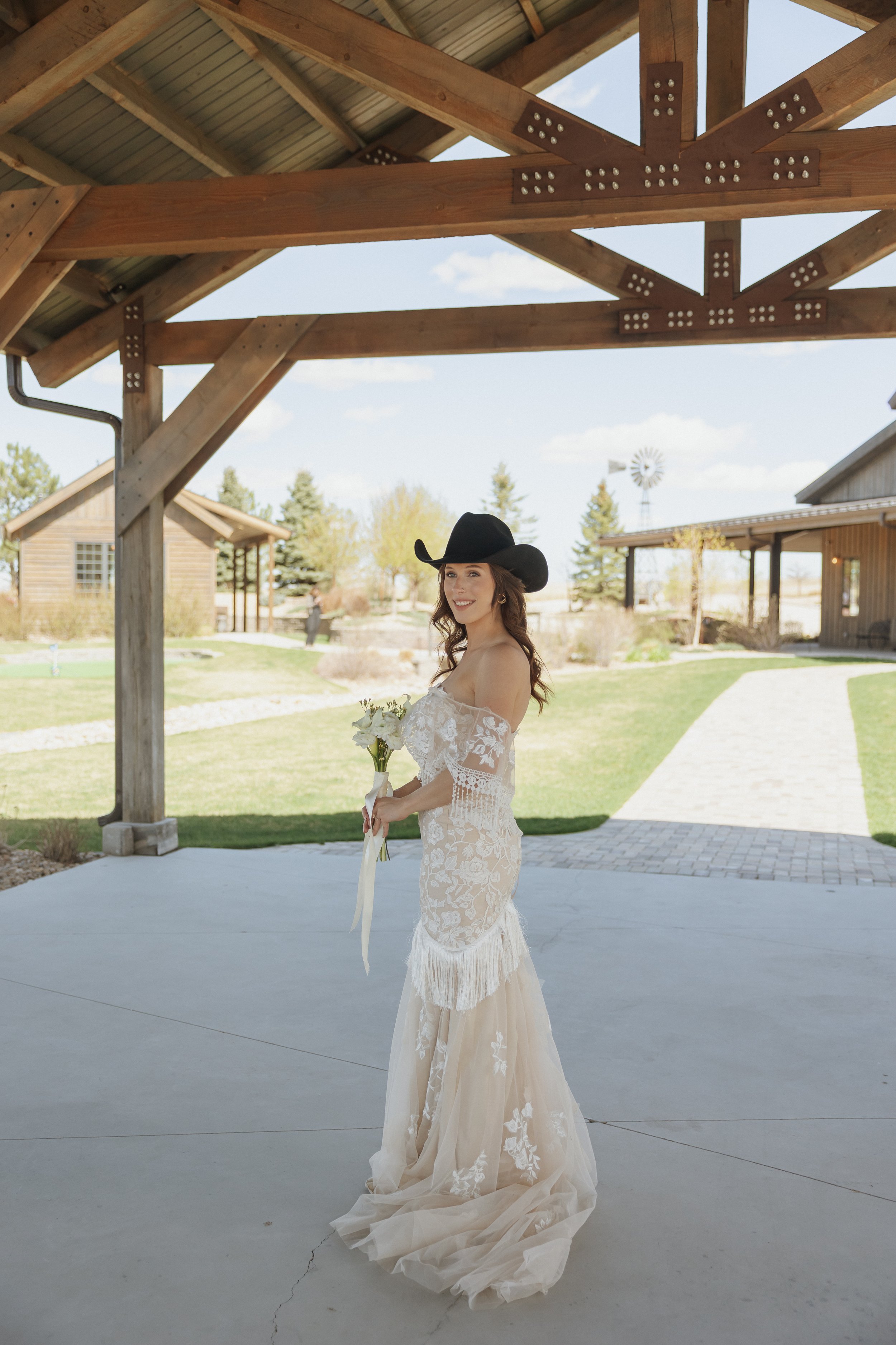 A woman in a white lace wedding dress and black cowboy hat stands outdoors holding a bouquet of flowers, smiling under a wooden pavilion with a rural landscape in the background.