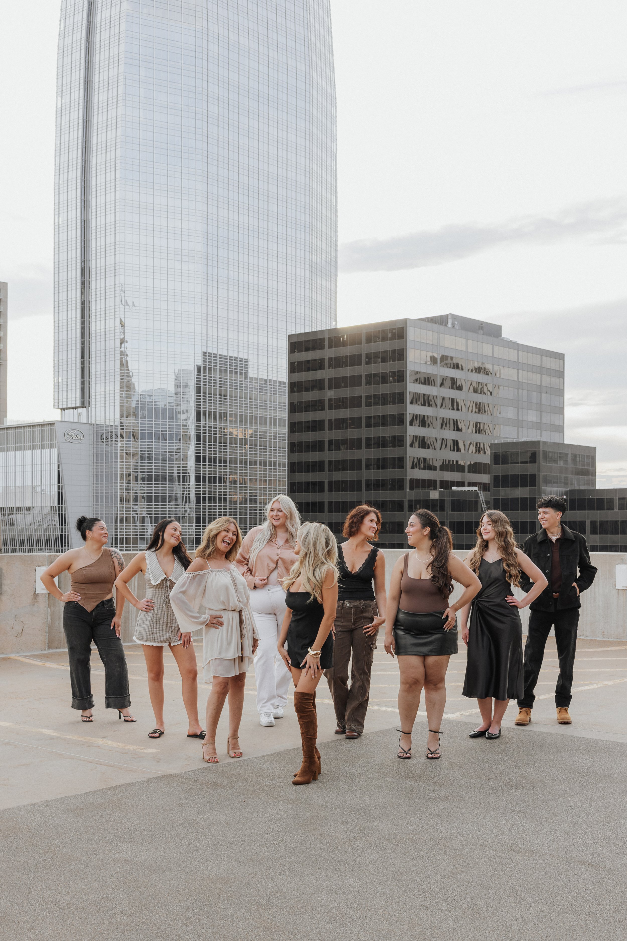 Group of women and one man standing and talking on a rooftop parking lot with tall office buildings in the background.
