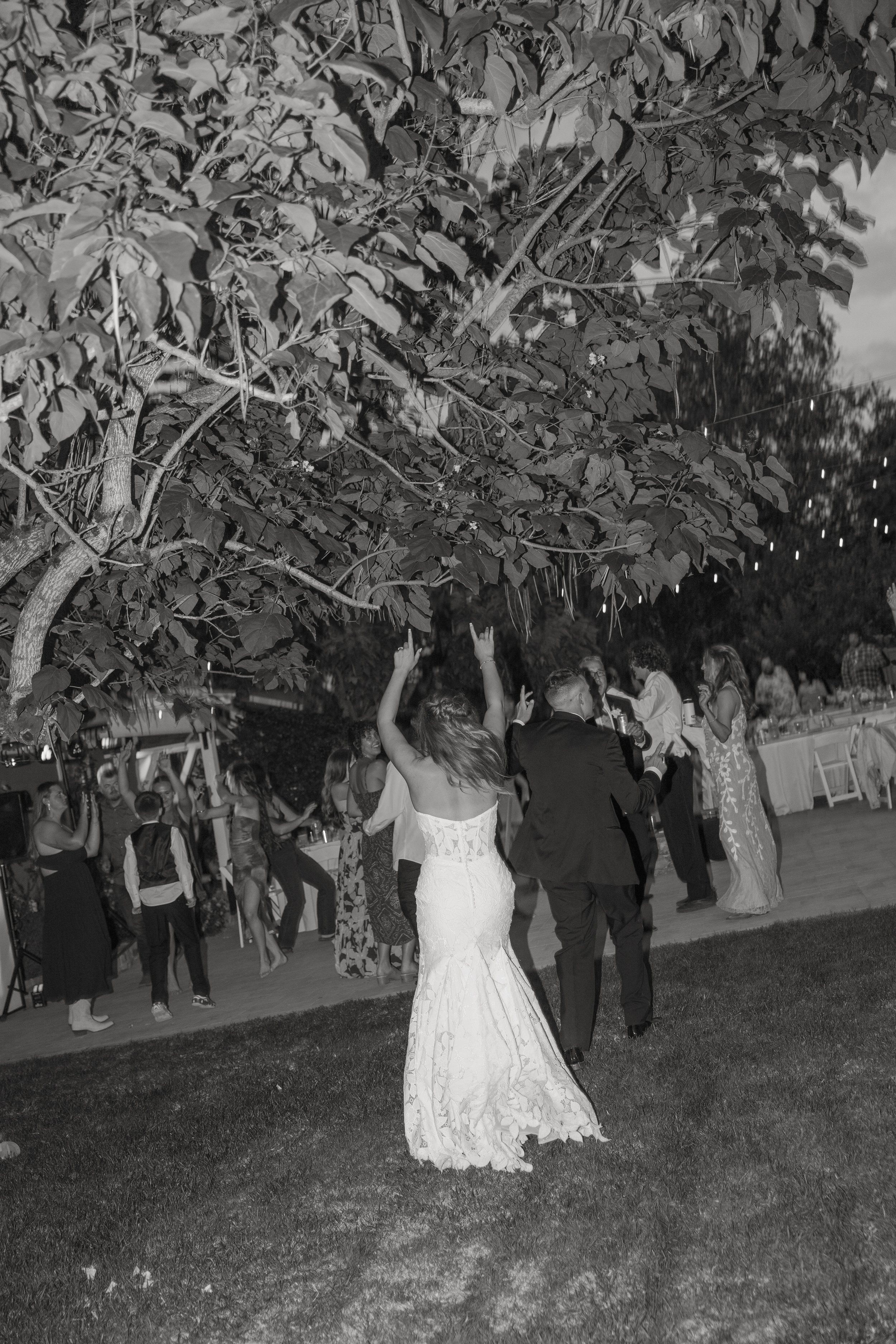 A black and white photograph capturing a lively outdoor party with people dancing and socializing. Prominent in the center is a woman in a white wedding gown, facing away and dancing. The scene is set against a backdrop of trees and a fence, with str