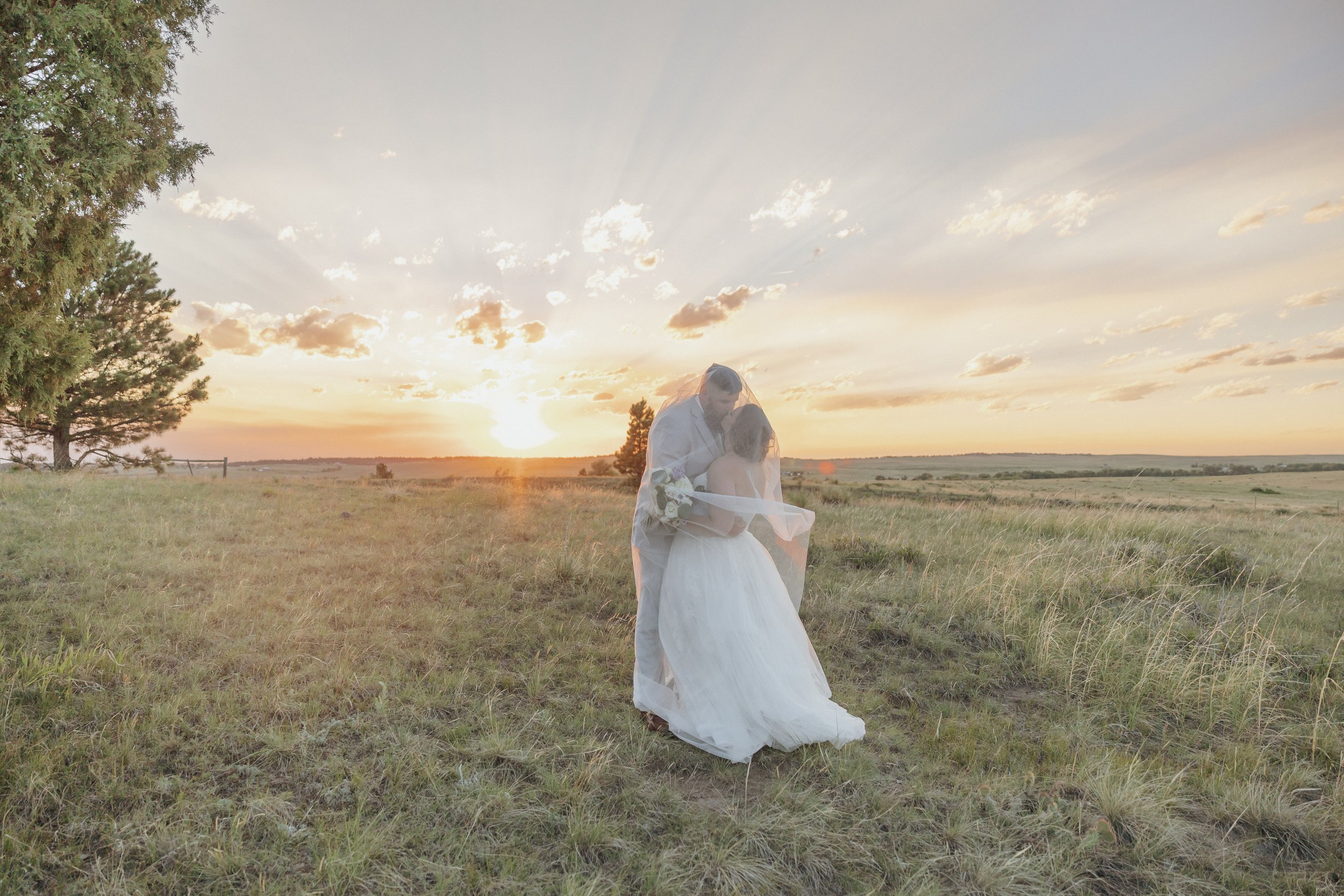 A bride and groom share an intimate kiss in an open grassy field at sunset. The bride wears a white wedding dress and veil, while the groom is dressed in light-colored formal attire. The sky is filled with soft clouds and the sun is low on the horizon, creating a warm, golden light.