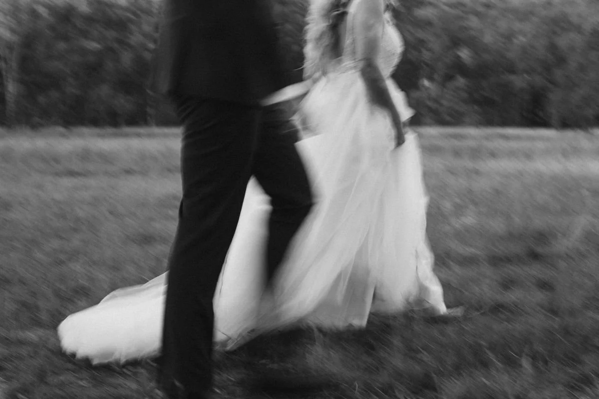 Person in black clothing walking on grass while holding a long, flowing light-colored fabric or dress outdoors.