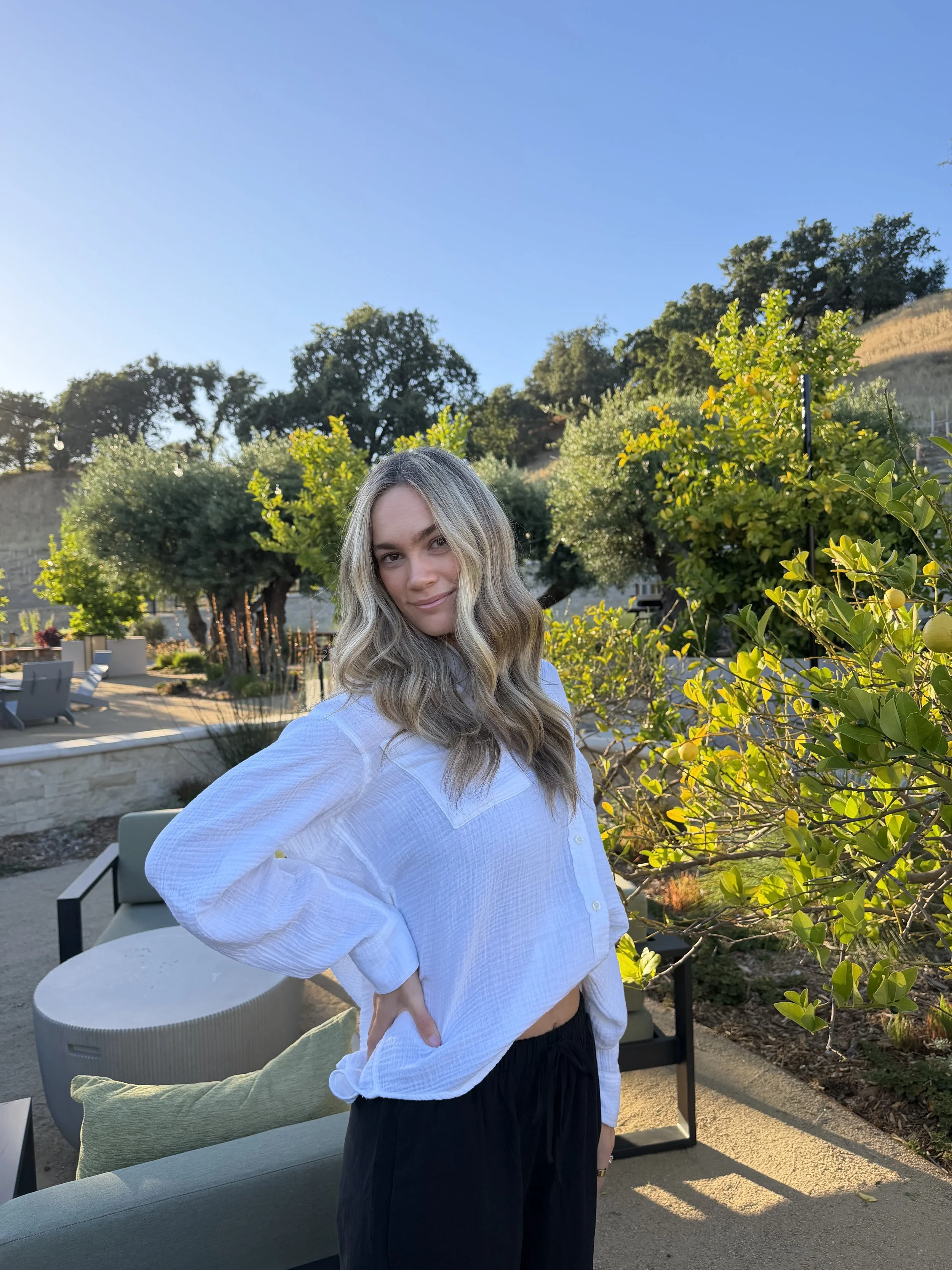 A woman with long wavy blonde hair wearing a white shirt and black pants standing outdoors in front of green bushes and trees, with a clear blue sky overhead.