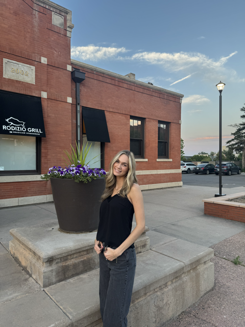 Young woman with blonde hair smiling in casual black top and jeans standing outside of a brick building with a black awning for a restaurant called Rodizio Grill, near a large planter with purple flowers, during sunset.