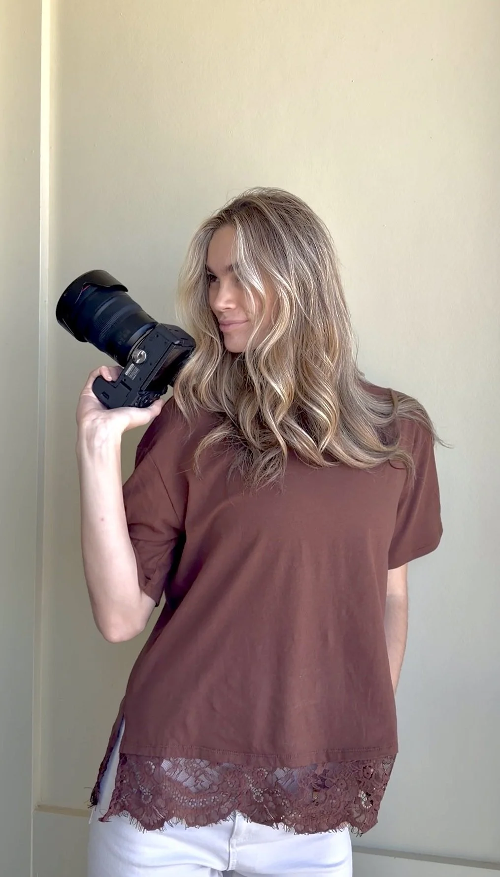 A woman with wavy blonde hair holding a professional camera, standing against a plain, light-colored wall.