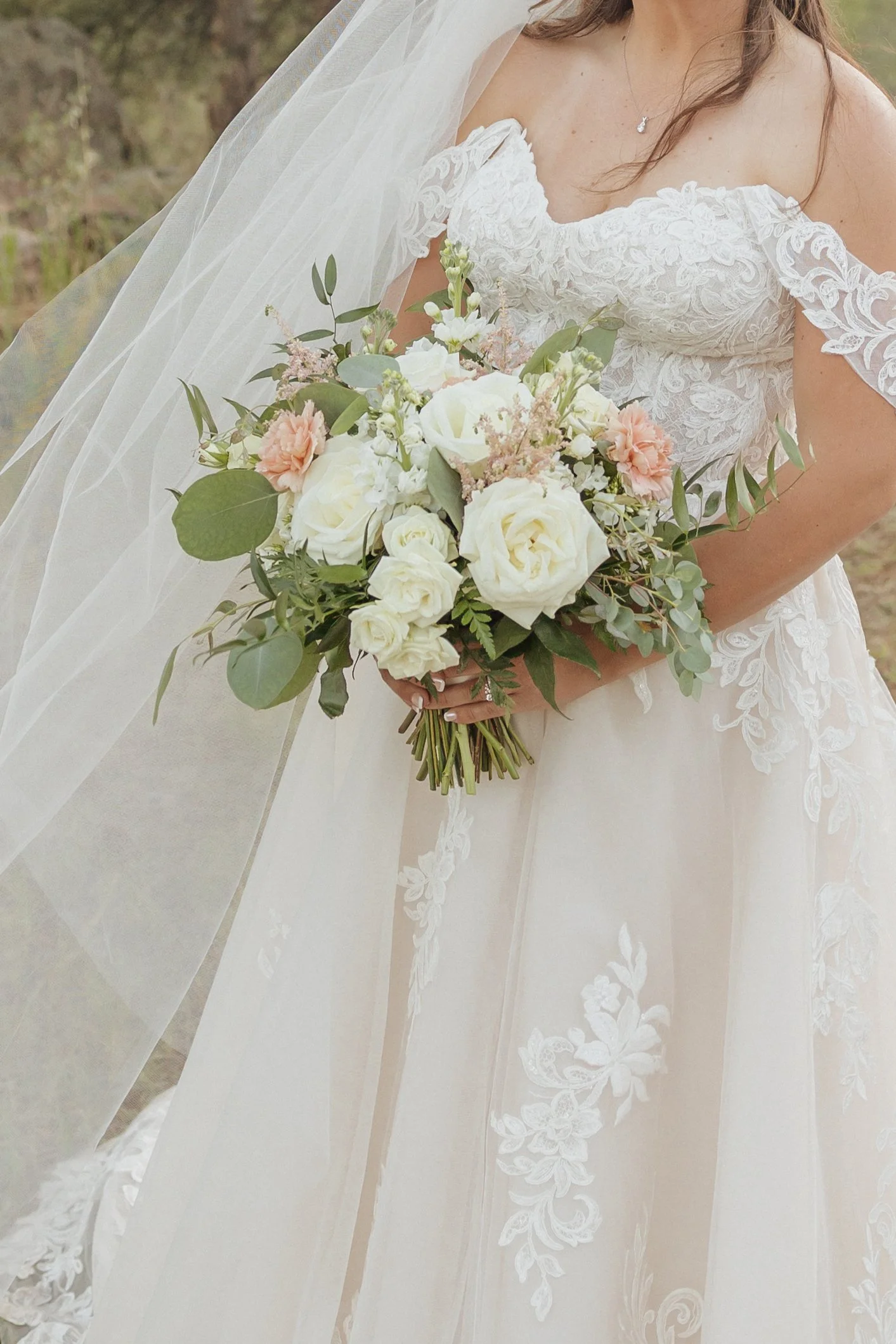 A bride in a white lace wedding dress holding a bouquet of white and pale pink roses, with greenery, standing outdoors.