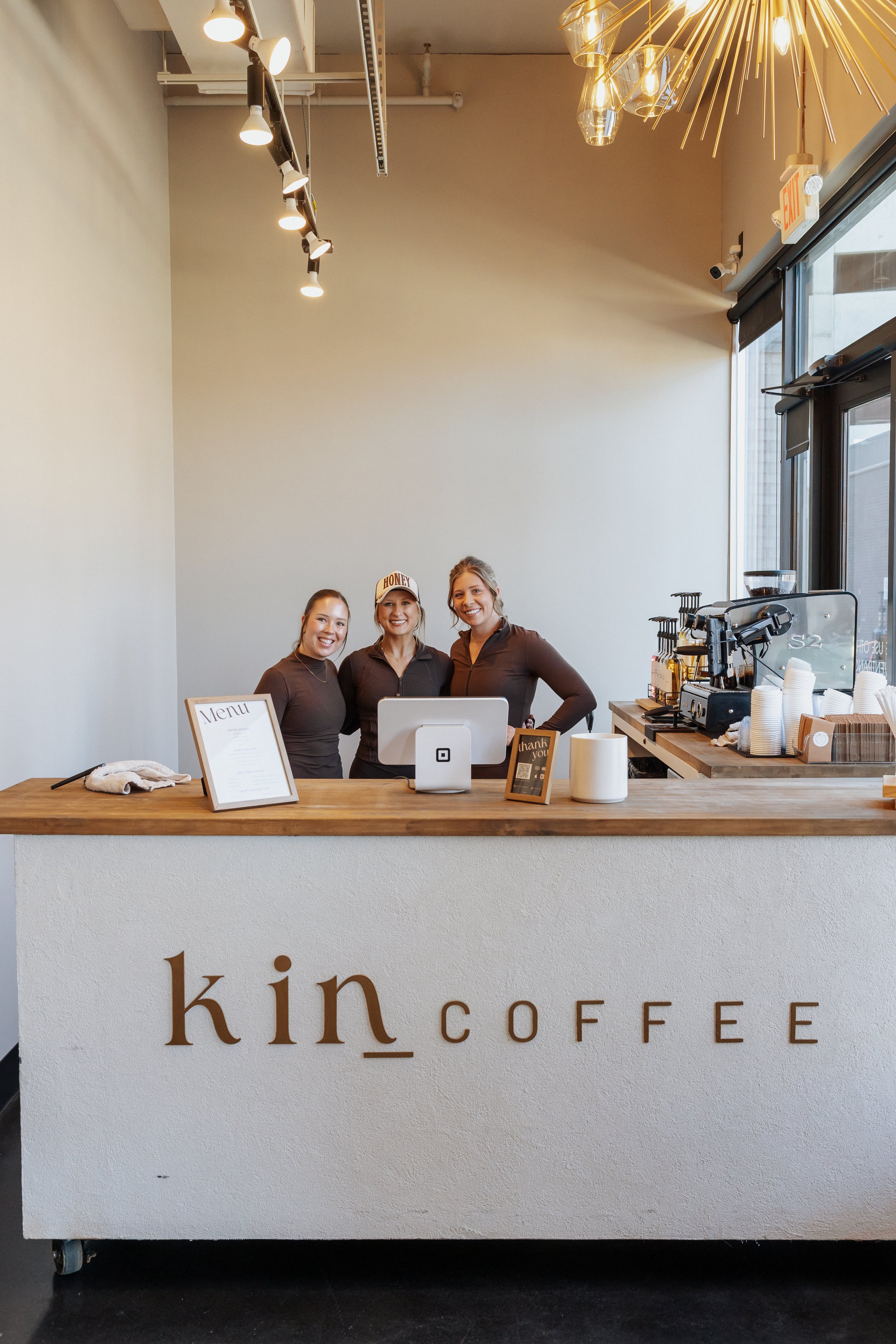 Three women standing behind a café counter smiling for the camera. The counter has a menu, a thank you card, and a cup. The background includes a coffee machine and a large window with an exit sign above.