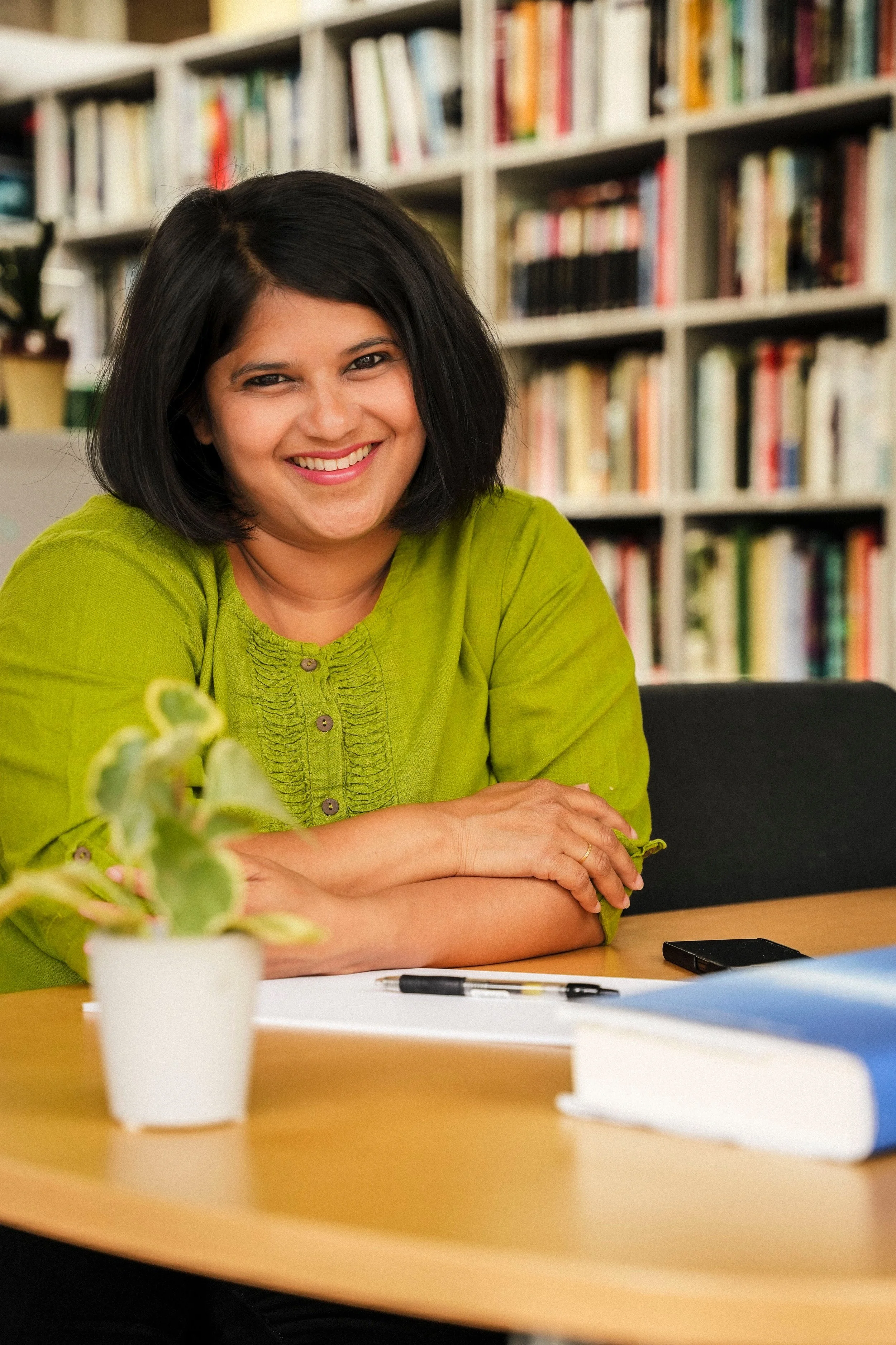 Woman smiling at a table in front of bookshelves, wearing a green top, with a plant and books in the foreground. Mansee Gupte