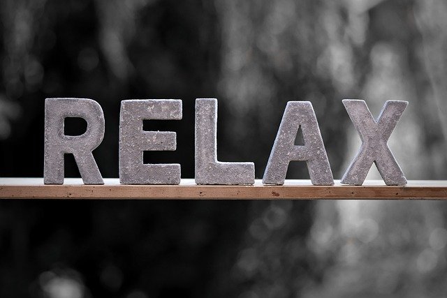Concrete letters spelling "RELAX" resting on a wooden shelf against a softly blurred dark background.