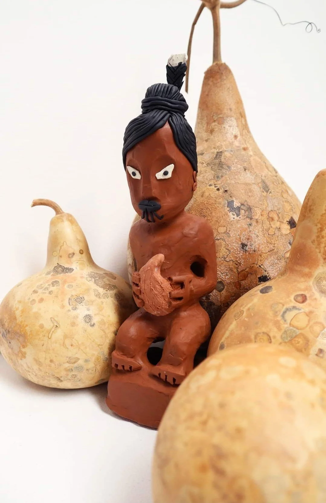 A clay sculpture of a person with black hair styled in a top knot, a moustache, and a beard, surrounded by several gourds.