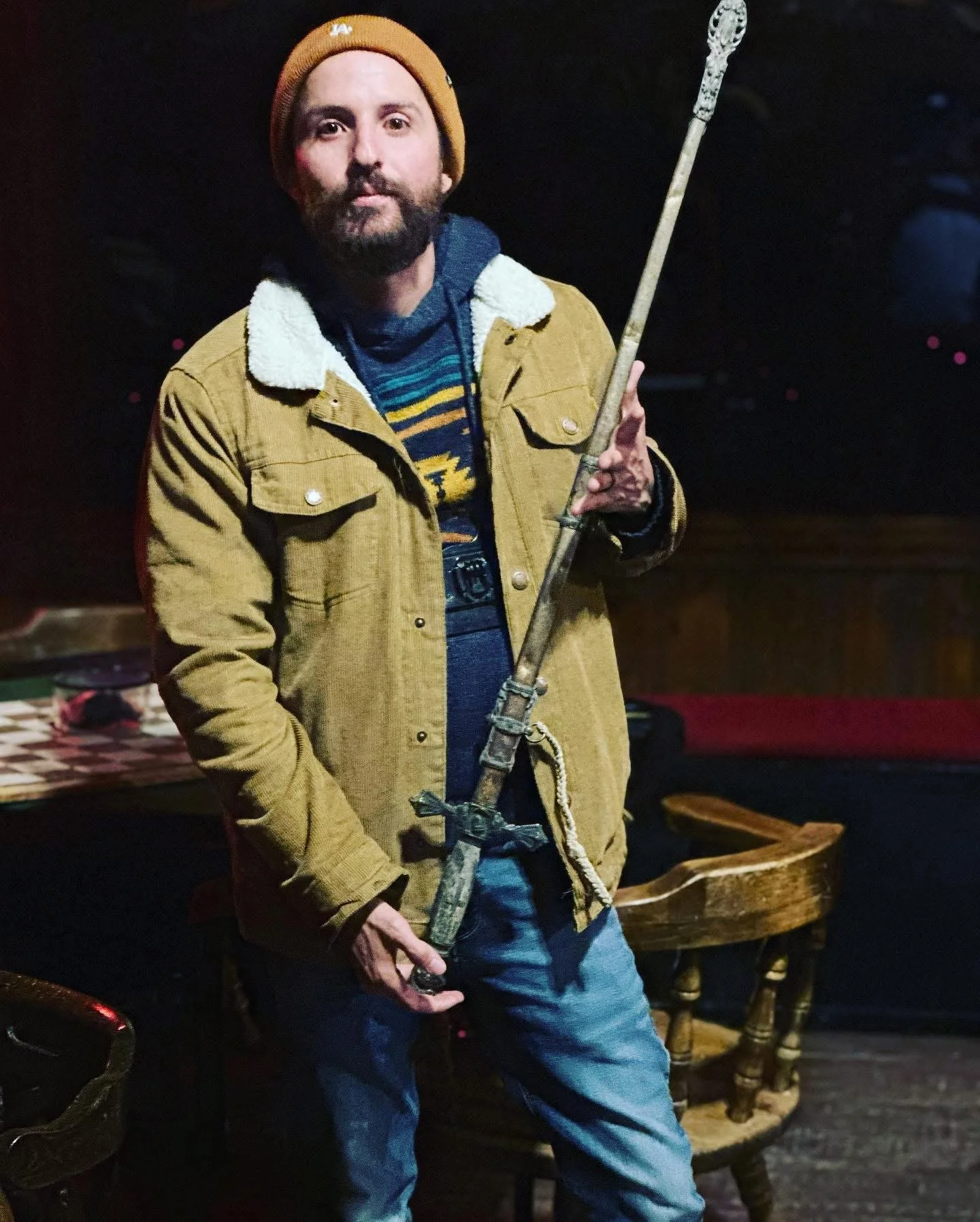 A person with a beard and mustache wearing a brown beanie and a tan jacket with a white collar, holding a staff or spear, standing in a dark room with wooden furniture and a checkered tablecloth in the background.