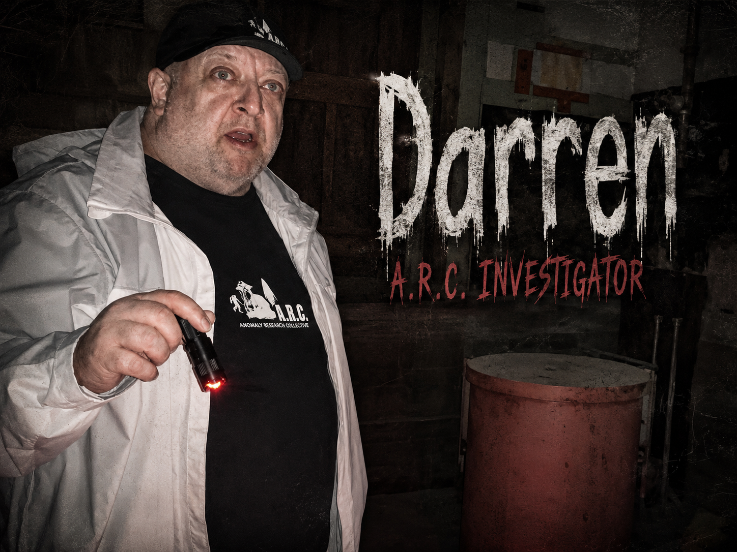 A man holding a small black flashlight in a dark basement, wearing a black cap and a black T-shirt with the logo 'A.R.C.' and the words 'Anomaly Research Collective'.