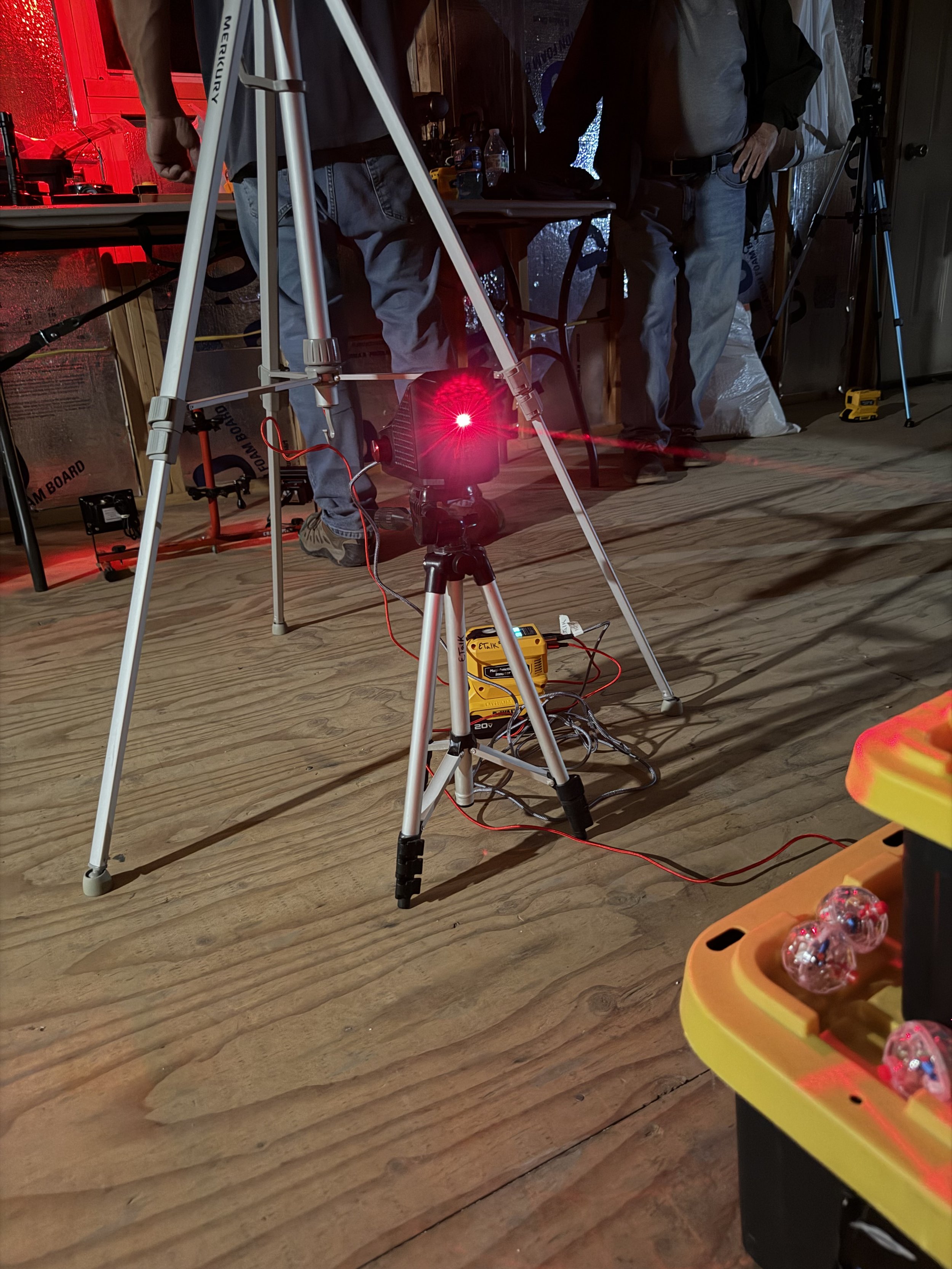 Indoor scene with a red laser grid emitted from a paranormal device on a tripod, research team ready to find the anomalies, a rem pod, a tripod, and a table with research equipment, including a yellow container with catballs.