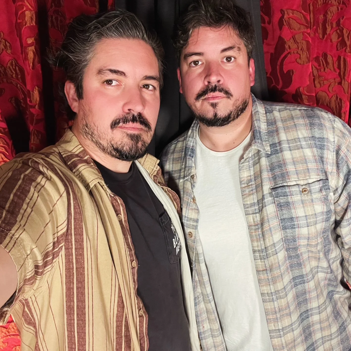 Two men standing close together, posing for a selfie in front of red patterned curtains.
