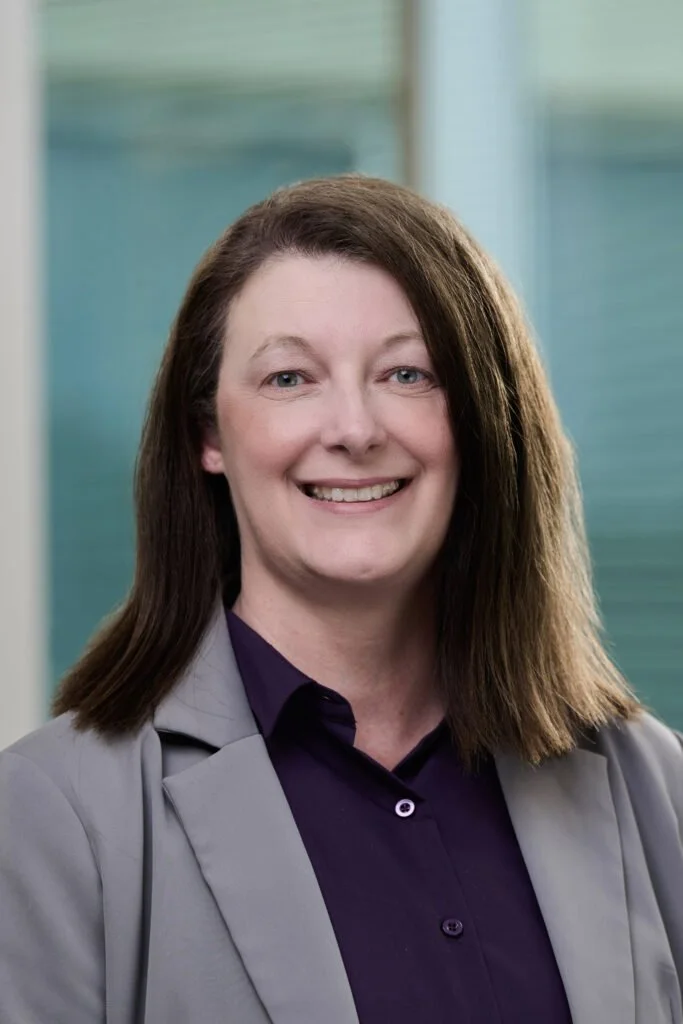 A woman with shoulder-length brown hair, wearing a gray blazer and dark purple shirt, smiling in front of a blurred blue background.