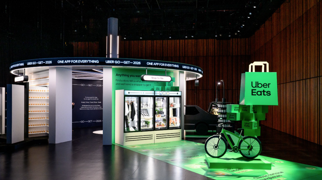 Uber Eats promotional display with a large green shopping bag and bicycle, a vending area, and digital screens inside a modern, wood-paneled room.