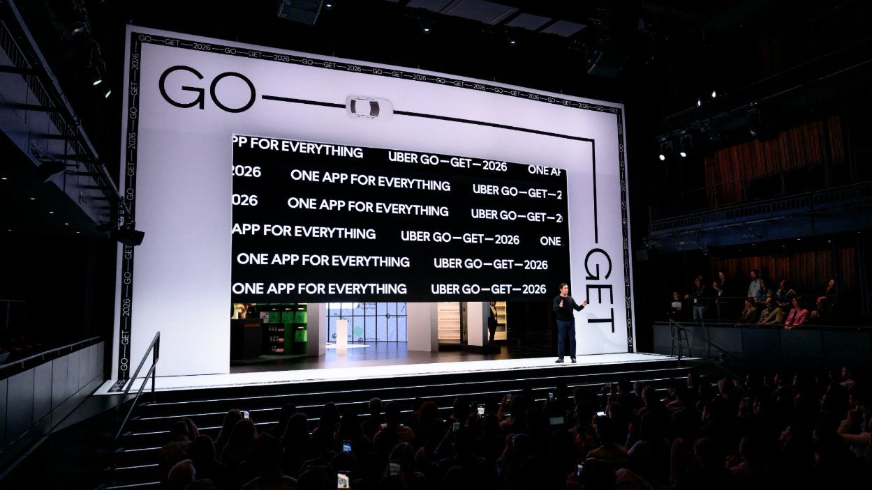 A speaker is standing on stage in front of a large screen that displays repetitive promotional text about Uber's app for 2026. The audience is seated and watching the presentation in a dark auditorium.