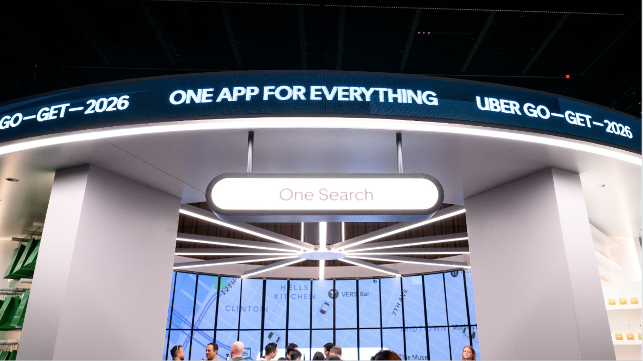 A digital display in an airport or shopping mall with promotional messages for Uber, a search app, and a tagline 'One app for everything'. People are visible sitting and walking below, against a large window showing a map and city streets outside.