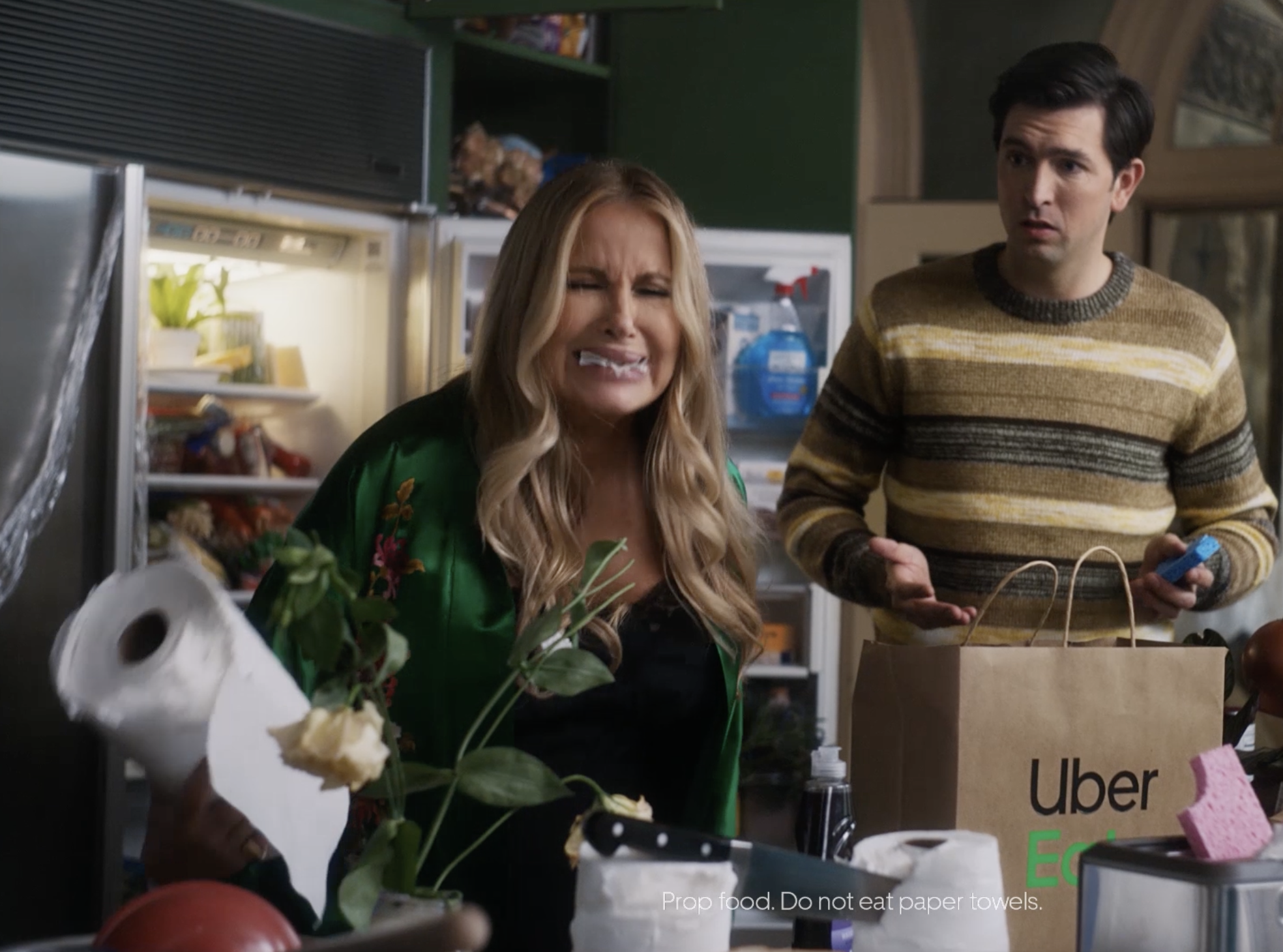 A woman with long blonde hair and a green floral dress is eating food with paper towels on her face, standing in a kitchen. A man with short dark hair, wearing a striped sweater, is standing next to her holding a smartphone. The refrigerator is open in the background. A cardboard box with Uber Eats logo is on the table, along with a pink sponge and paper towels with a note that says, "Prop food. Do not eat paper towels."