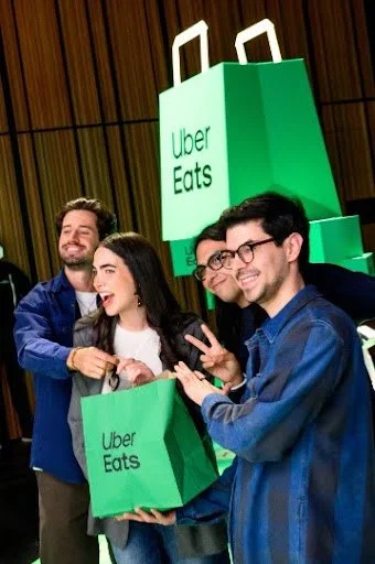 Four people smiling and posing with Uber Eats shopping bags in front of a large Uber Eats logo.