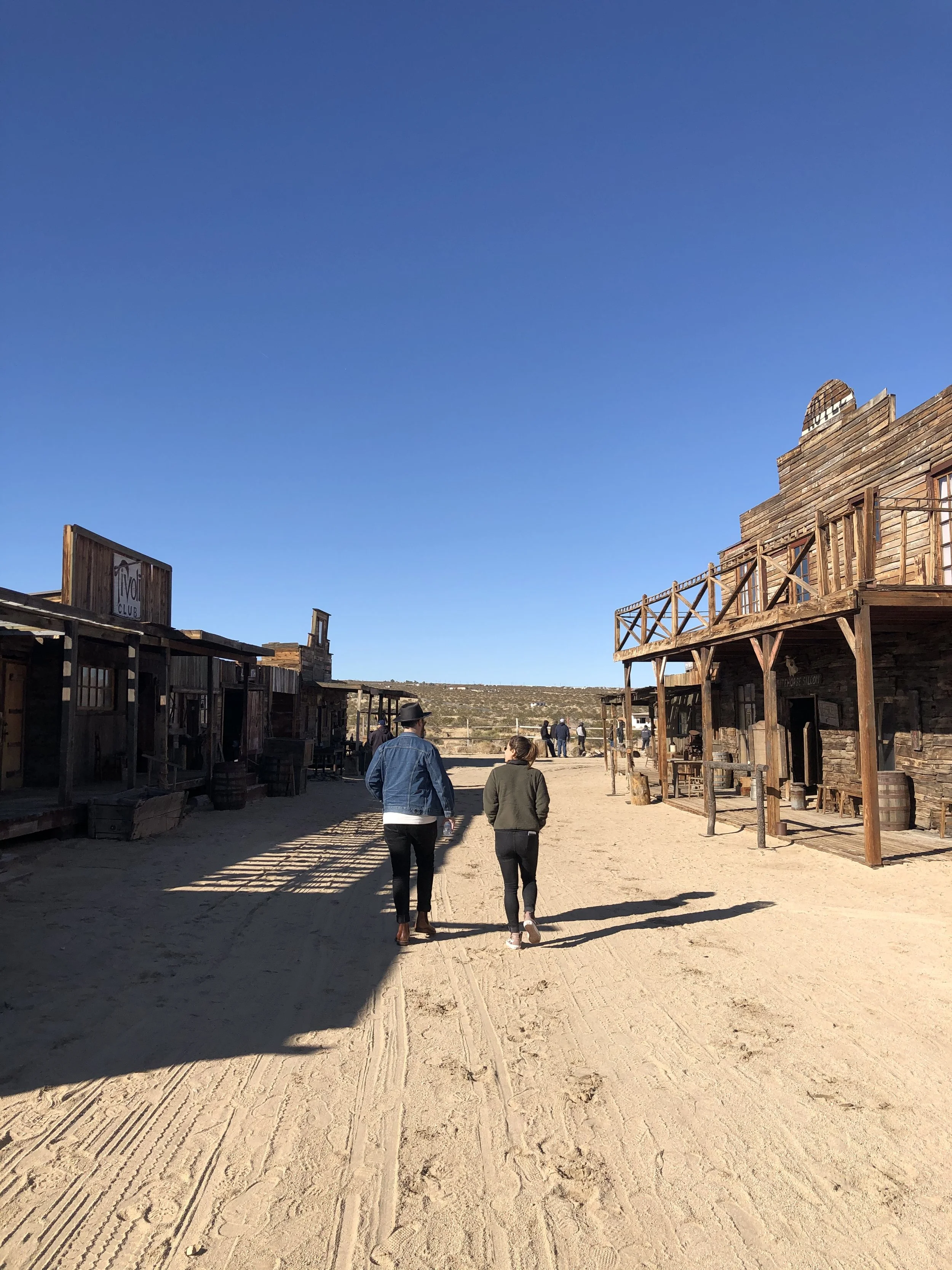 A Western-themed town street with wooden buildings on either side, a clear blue sky, and people walking away from the camera.