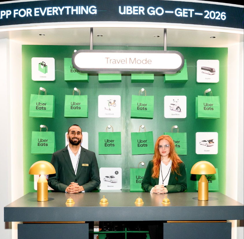 Two people standing behind a black table with yellow lamps, in front of a green wall decorated with Uber Eats bags and icons of various transportation modes, and a sign that says 'Travel Mode'.