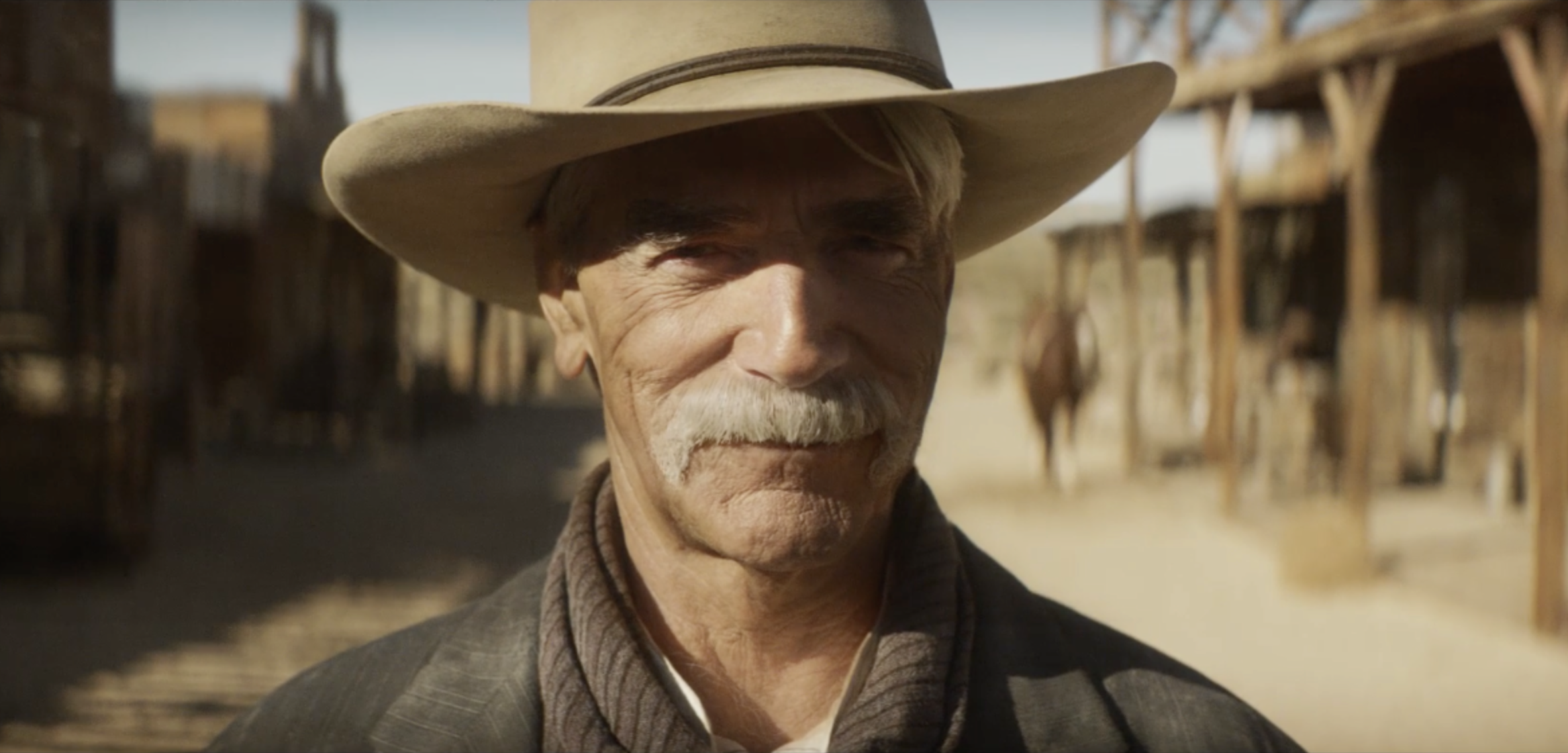 Close-up of an elderly man with a white mustache and wearing a cowboy hat, standing outdoors in a desert-like environment with wooden structures in the background.