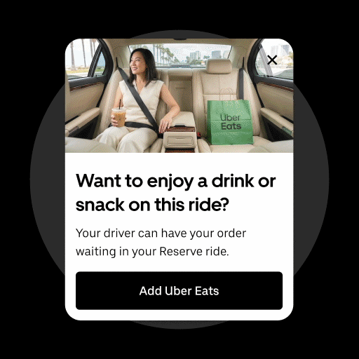 Woman sitting in the backseat of a car with a seatbelt on, holding a drink, and an Uber Eats bag placed beside her. Car interior visible with windows showing a cityscape outside.