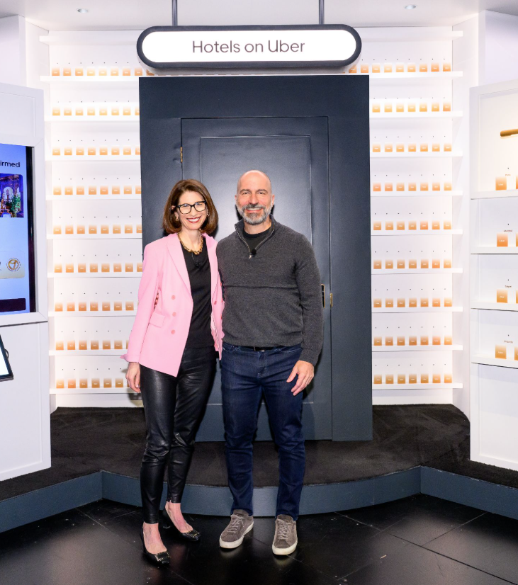 Two people, a woman wearing a pink blazer and black leather pants, and a man in a gray sweater and jeans, standing together inside a display area with shelves of orange boxes labeled 'Hotels on Uber.'