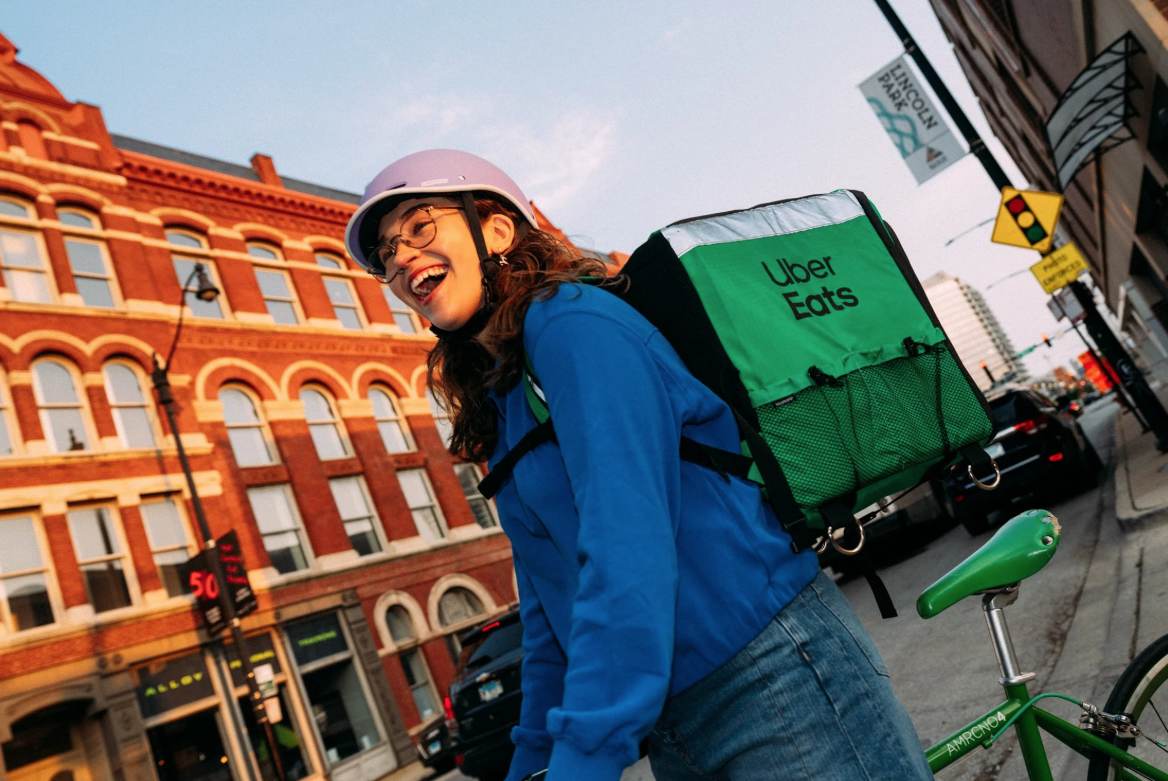 A smiling woman wearing a purple helmet and glasses with a blue jacket and jeans, standing next to a green Uber Eats delivery backpack, with a green bicycle.