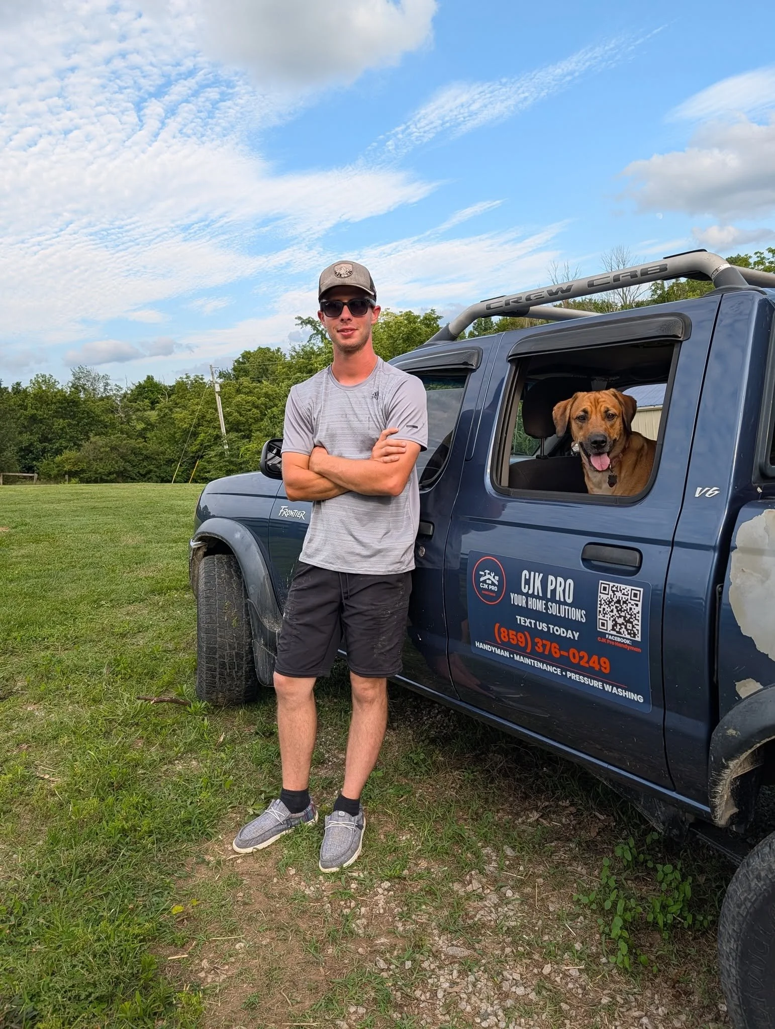 A young man wearing sunglasses, a cap, a gray T-shirt, and shorts, standing outdoors next to a blue pickup truck with a dog in the open window, against a scenic background of trees and a blue sky with clouds.