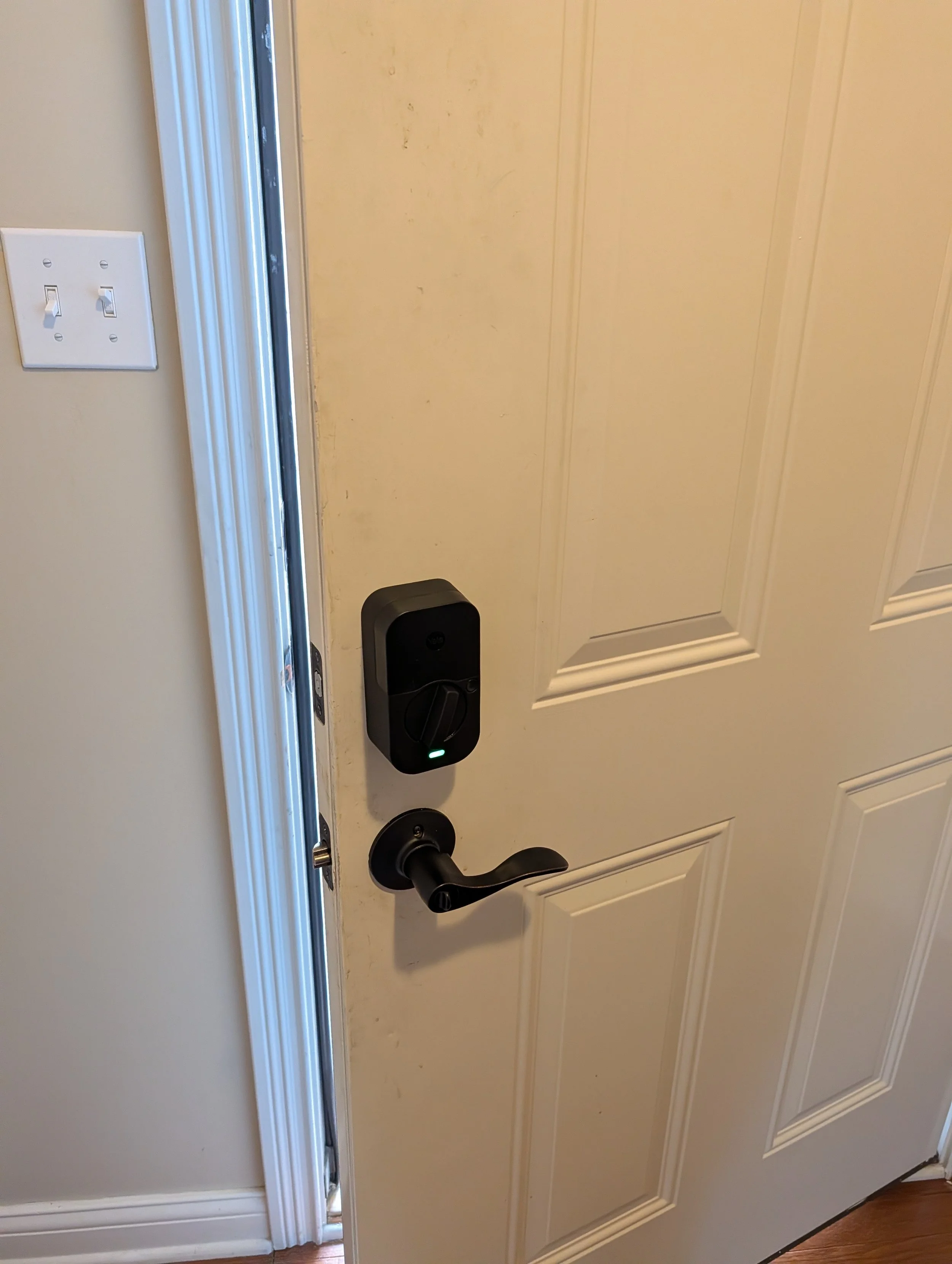A white front door with a black handle, a black electronic lock, and a black device on the ground near the door. There is a partial view of a light switch on the wall to the left of the door, and the door reveals a sliding glass door and a doorway leading outside.