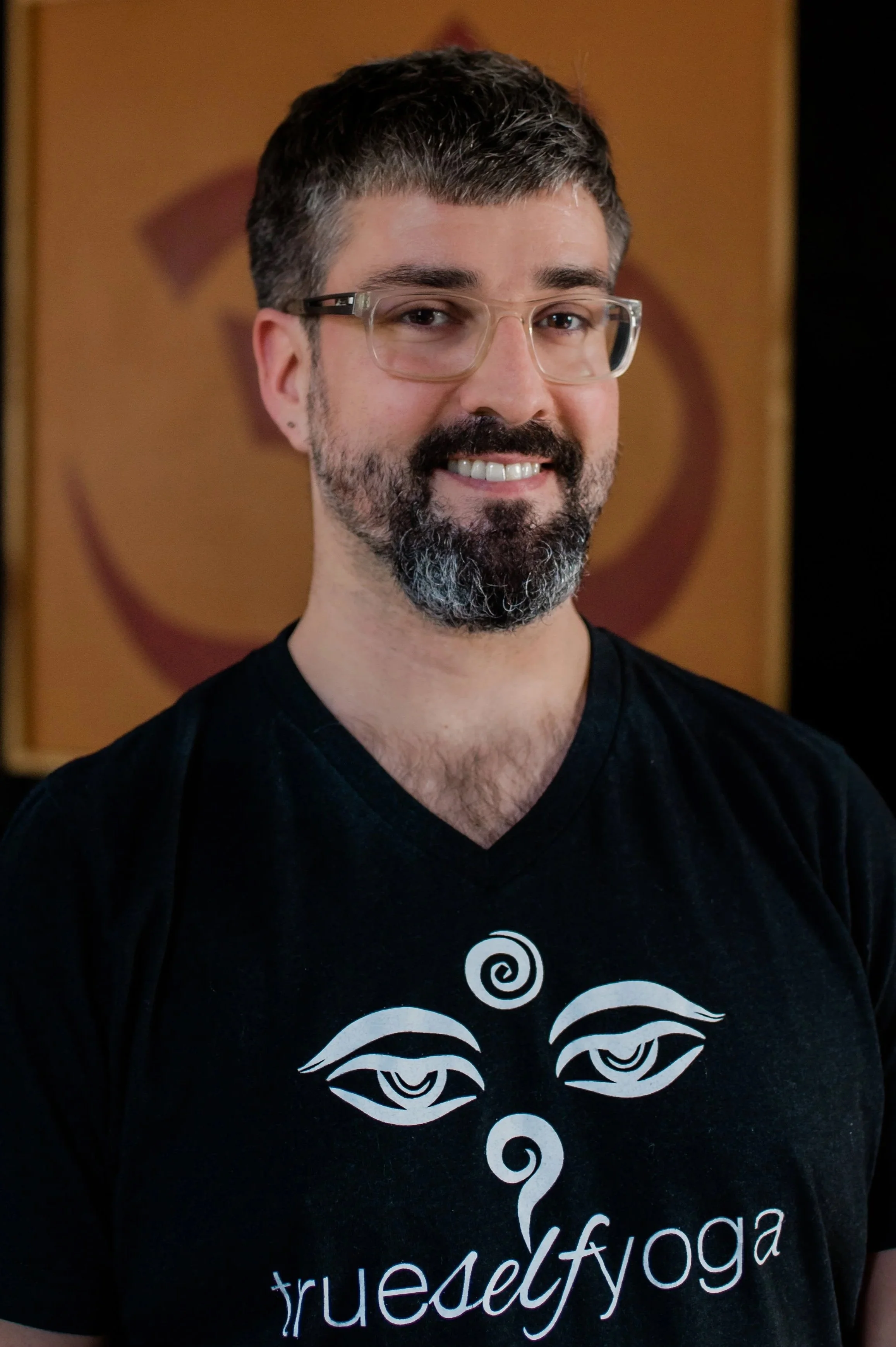 A man with glasses and a beard smiling, wearing a black T-shirt with a white graphic and text, standing in front of a brown background.