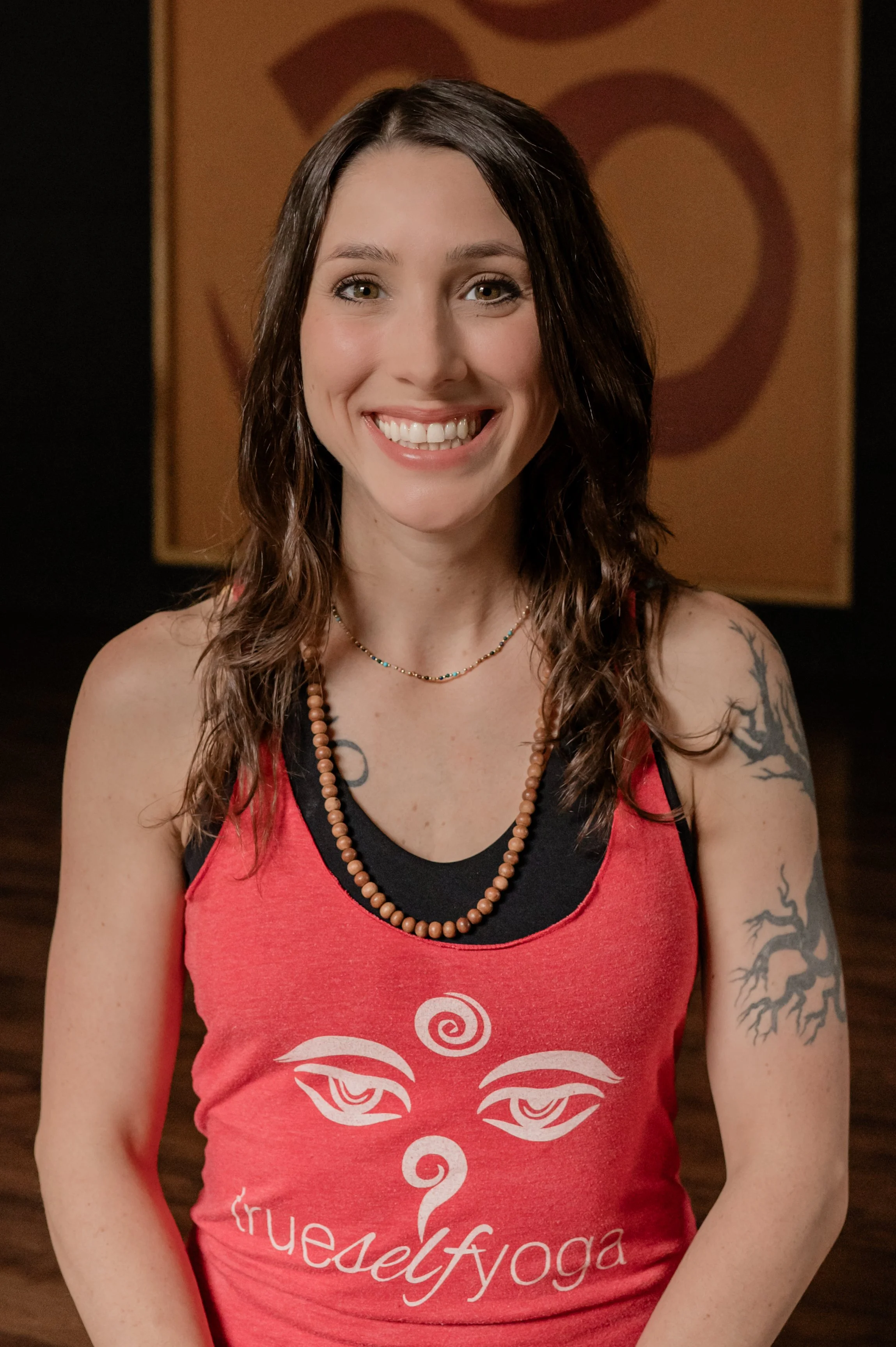 A young woman with long brown curly hair, smiling with braces, wearing a red tank top with white design and text, black trim, a beaded necklace, and a tattoo on her left upper arm. The background features a warm-toned cloth with a circular symbol.