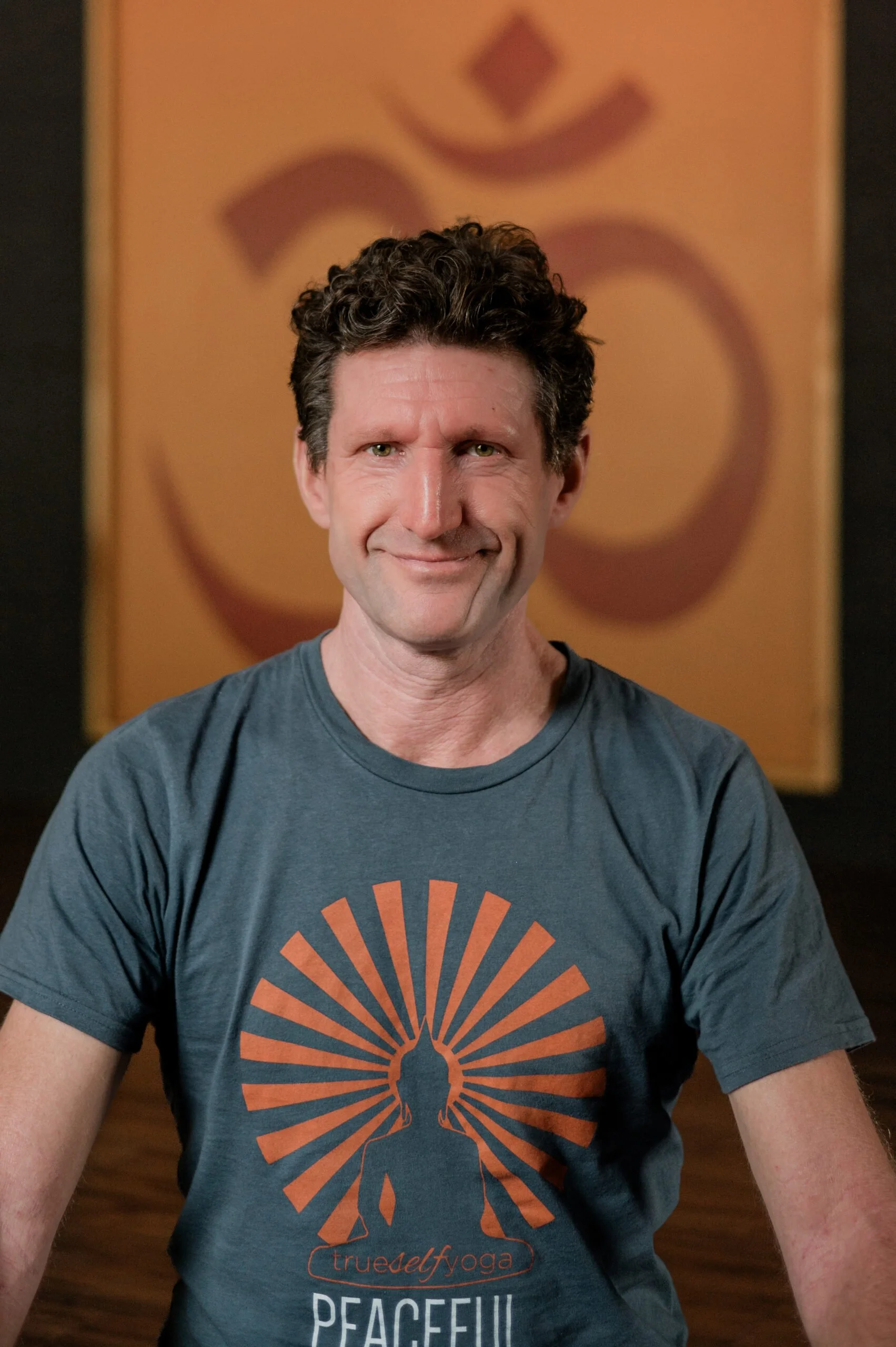 A man with curly dark hair and a light smile, wearing a gray T-shirt with an orange and blue logo and the words 'true yoga' and 'peaceful,' sits in front of a blurred background featuring a large orange poster with a black symbol or letter.