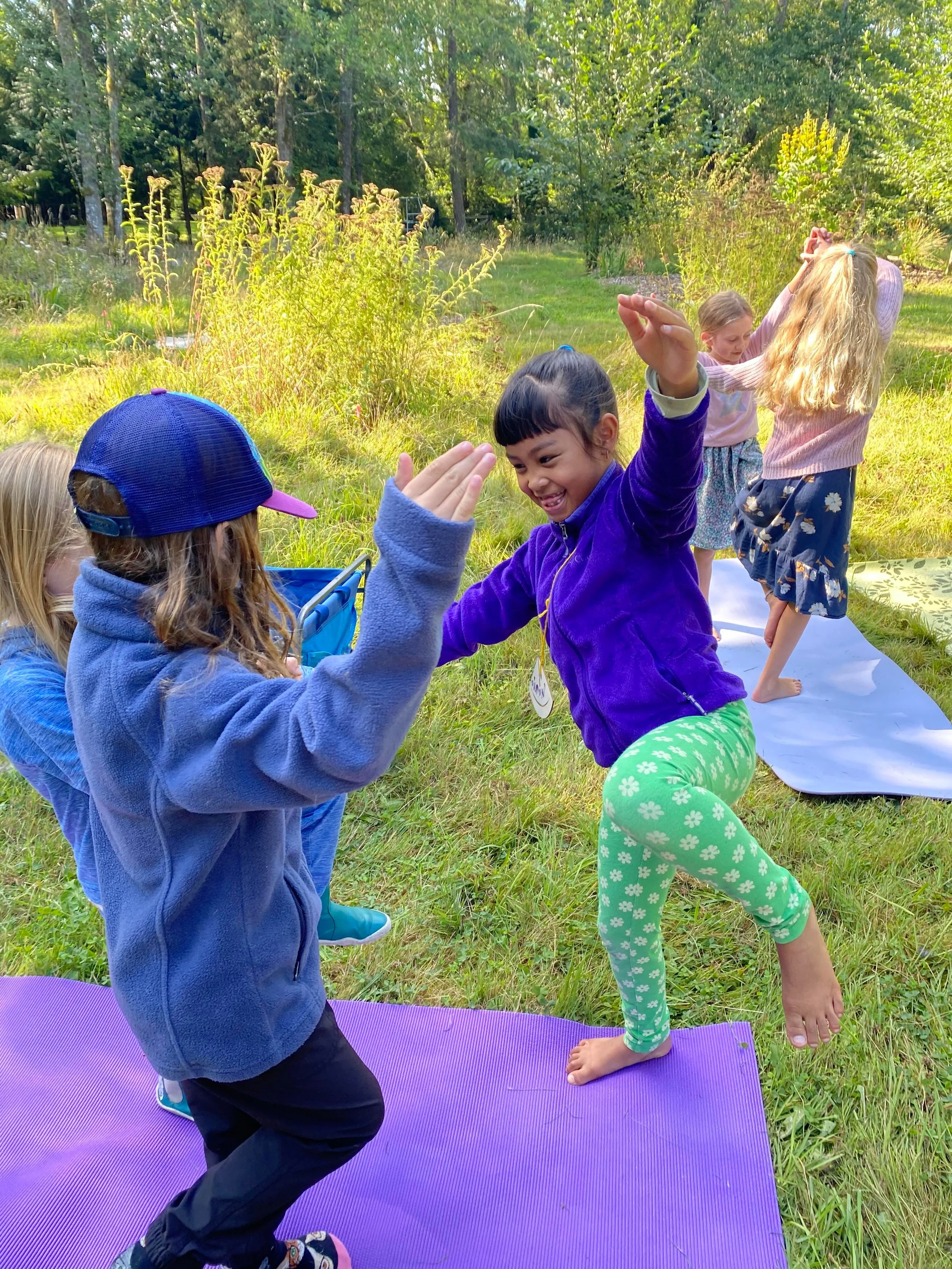 Group of children practicing yoga outside on purple mats in a green, wooded area.