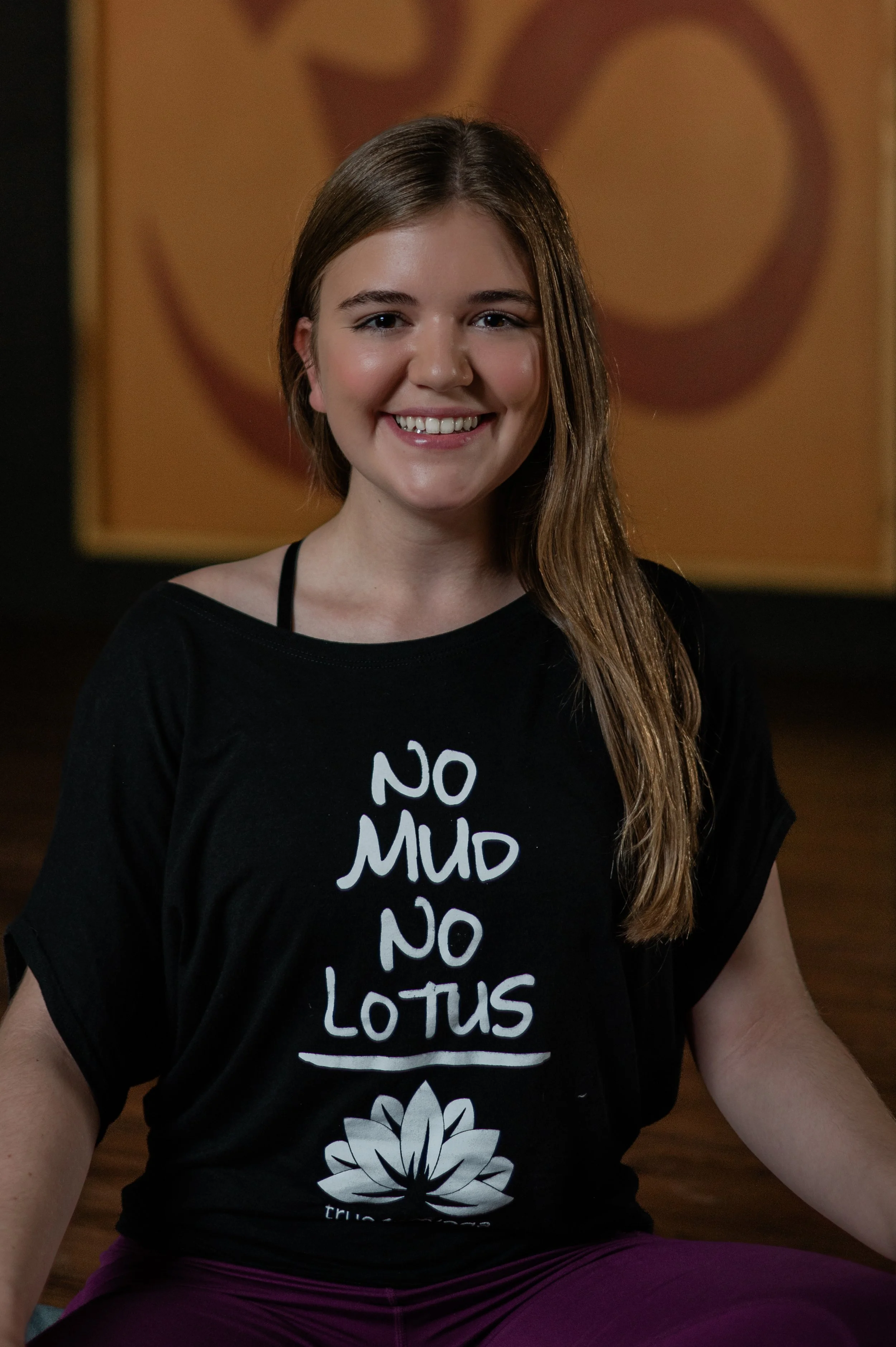 A young woman with long brown hair, smiling, wearing a black T-shirt with white text that reads 'NO MUD NO LOTUS' and a lotus flower graphic, sitting in a yoga pose in a studio with a heart-shaped wall art in the background.