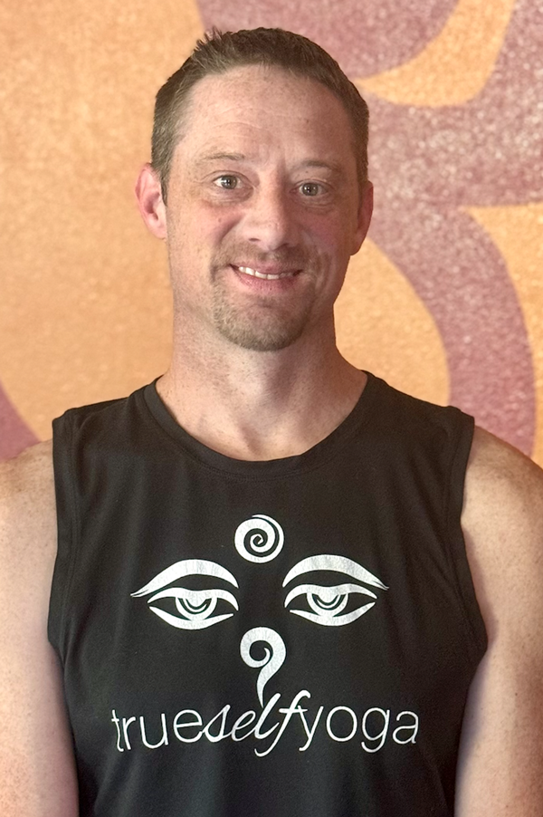 A man with short brown hair and a light beard, wearing a black sleeveless t-shirt with a yin-yang symbol and stylized eyes and the text 'trueselfyoga', standing in front of a patterned orange and purple background.