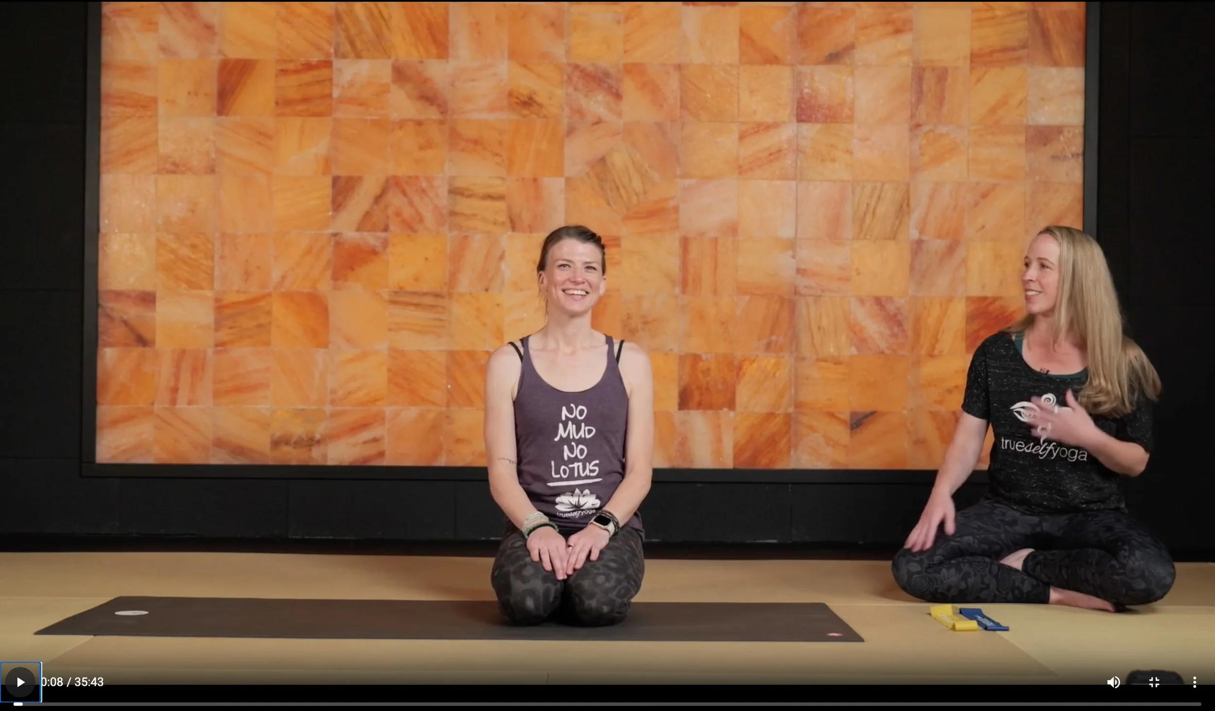 Two women in yoga attire sitting on a yoga mat, with a wooden wall background, one smiling and the other gesturing while talking.