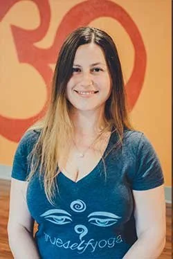 A young woman with long, light brown hair, smiling, wearing a navy blue t-shirt with a yoga-themed design, standing in front of a red background with orange and red patterns.