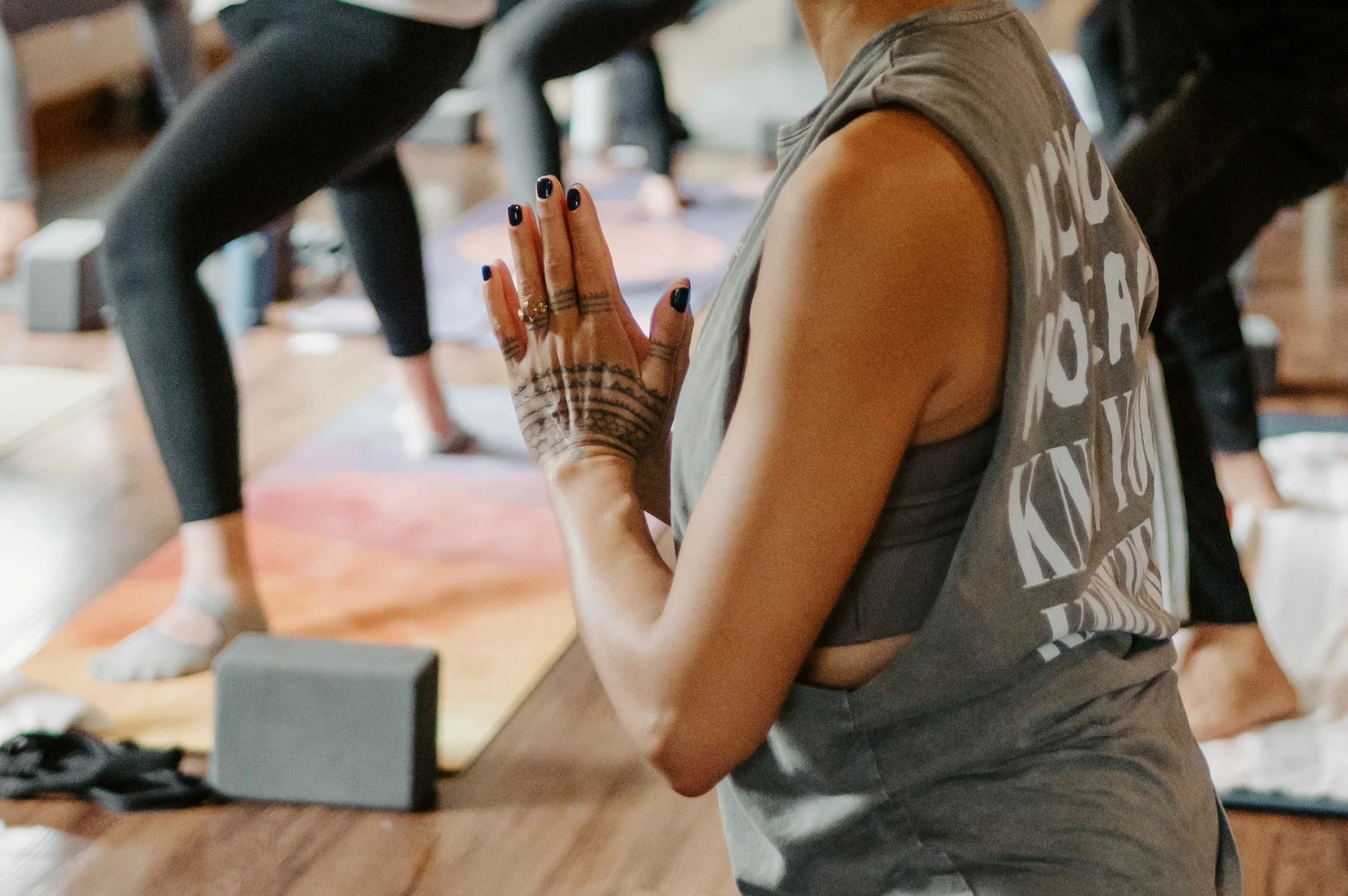 A woman in a sleeveless shirt with tattoos on her hand practicing yoga or meditation with her hands pressed together in a prayer position at a wooden table in a room with other people in the background.