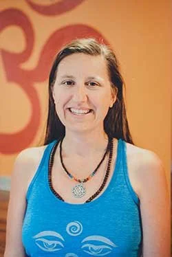 A woman with long dark hair wearing a blue tank top and multiple necklaces, smiling in front of an orange abstract background.
