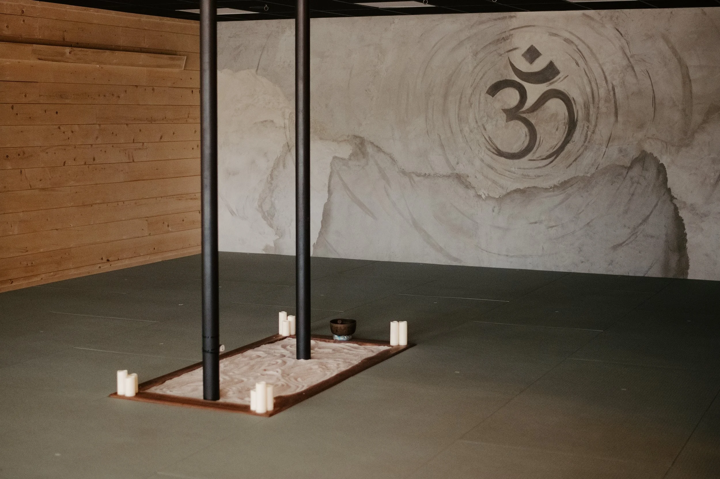 The interior of a minimalist room with a large wall featuring a black and white spiral galaxy mural. There are black metal poles, a small platform with white candles, a brown bowl, and a pinecone on a mat.