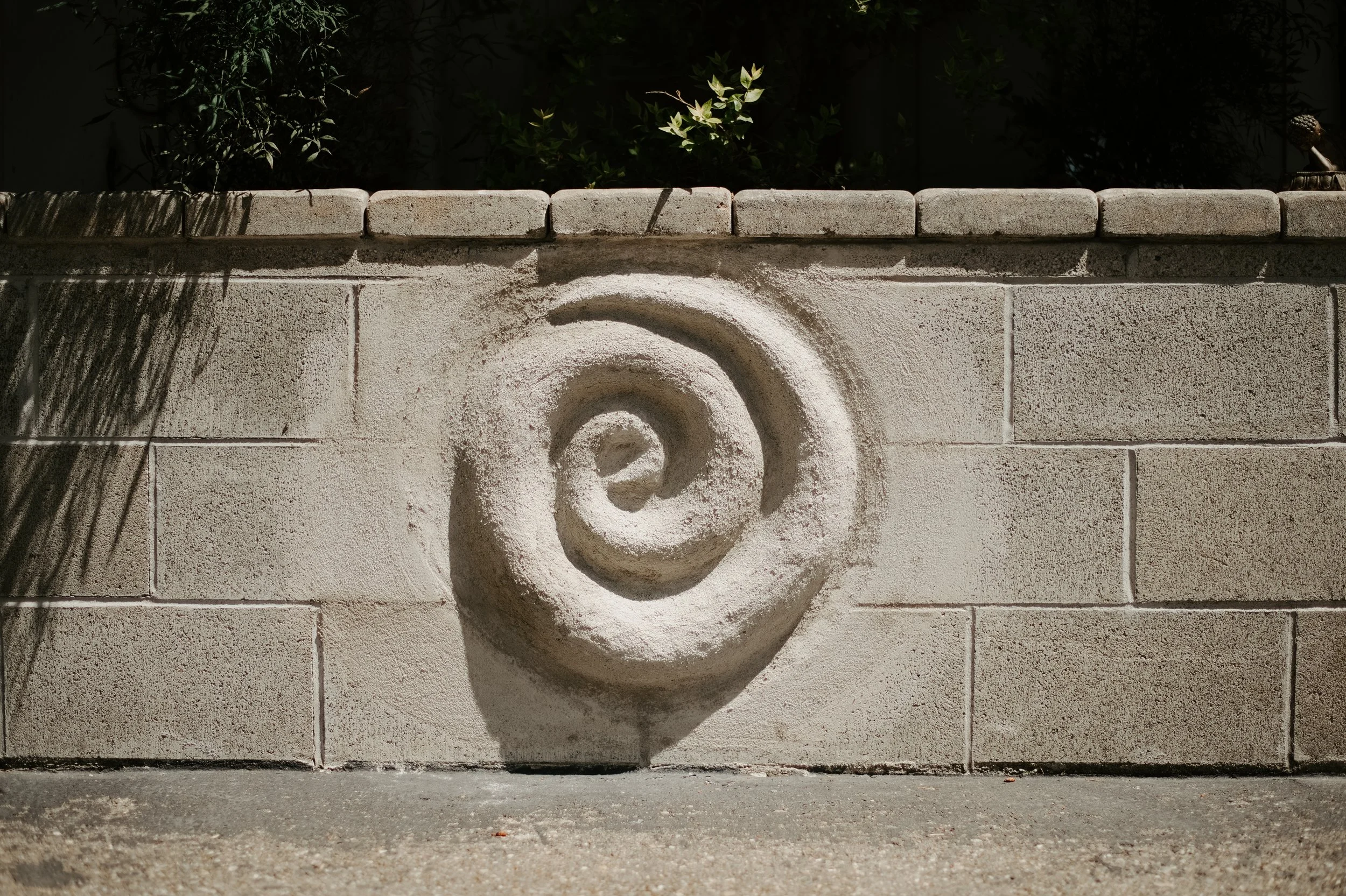 Close-up view of a textured, light-colored cement block wall with a spiral design carved into the lower middle section. Shadows of tree branches fall across the top part of the wall, and some greenery is visible behind it. The ground in front is concrete.