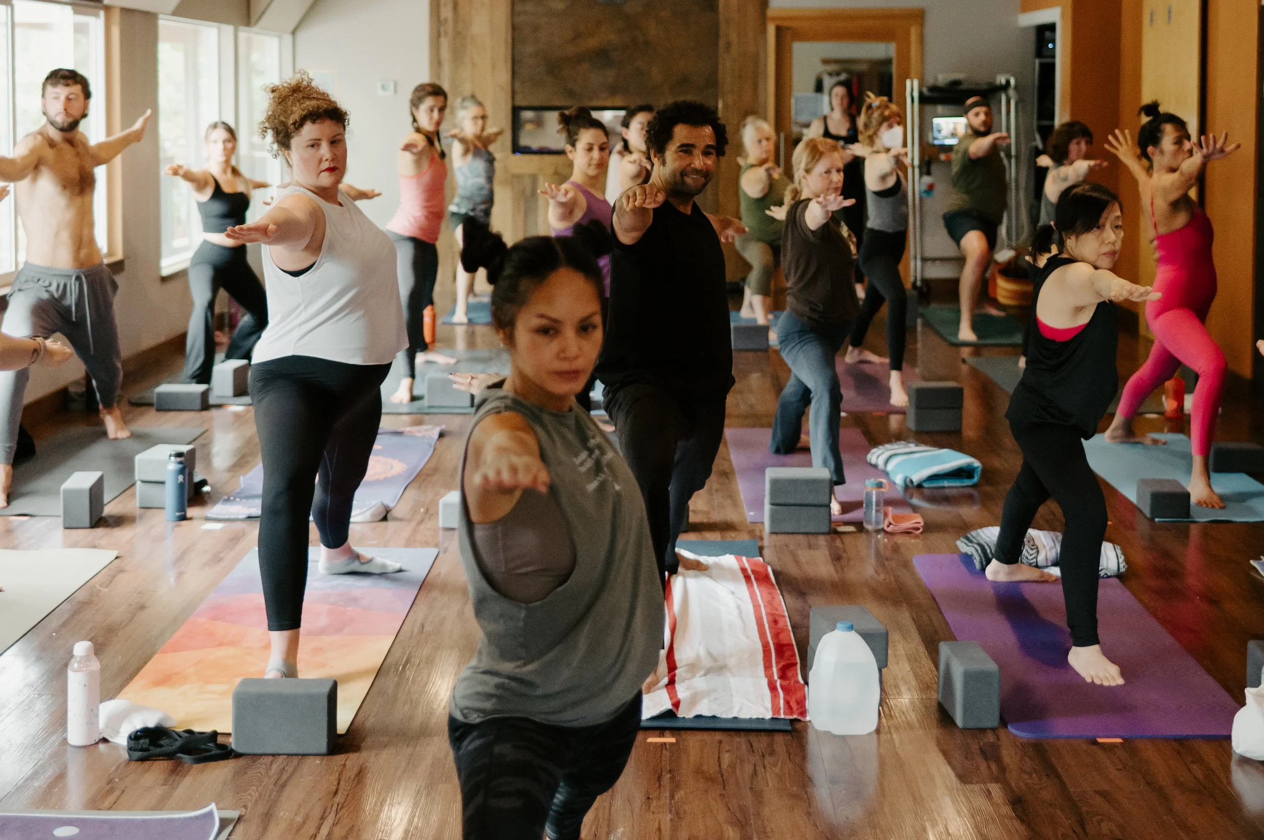 A diverse group of people participating in a yoga or fitness class indoors, performing a balancing pose on yoga mats with props like blocks and water bottles nearby.