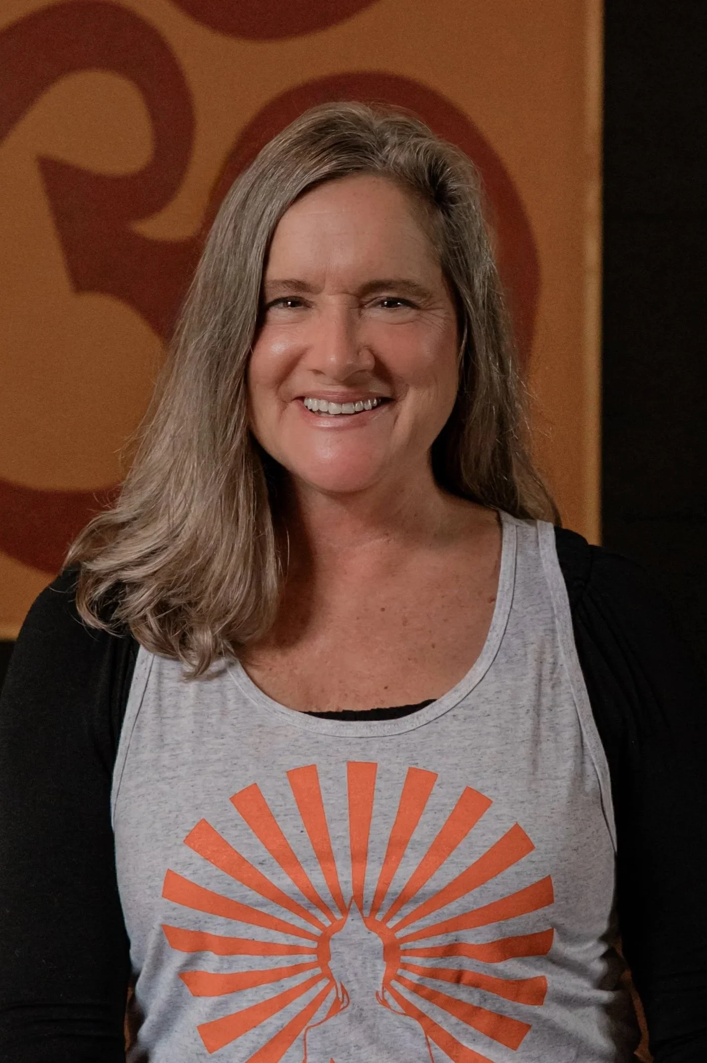 A woman with shoulder-length gray hair and a big smile wearing a gray tank top with an orange sunburst and tree design, standing in front of a brown background with abstract shapes.