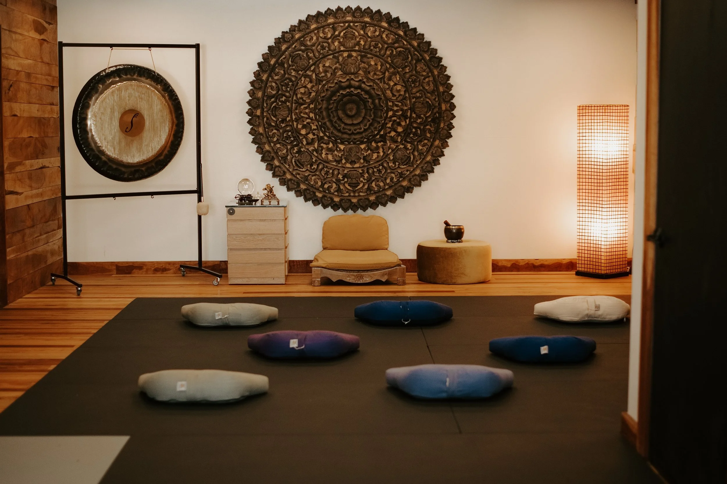 Inside a meditation or yoga studio with a black mat on the wooden floor, several round cushions in white, purple, and blue colors arranged for seating. The back wall features a large ornate wooden circular carving, a small beige cushion and a small table with spiritual or decorative items, and a standing lamp with a grid pattern shade emitting warm light.
