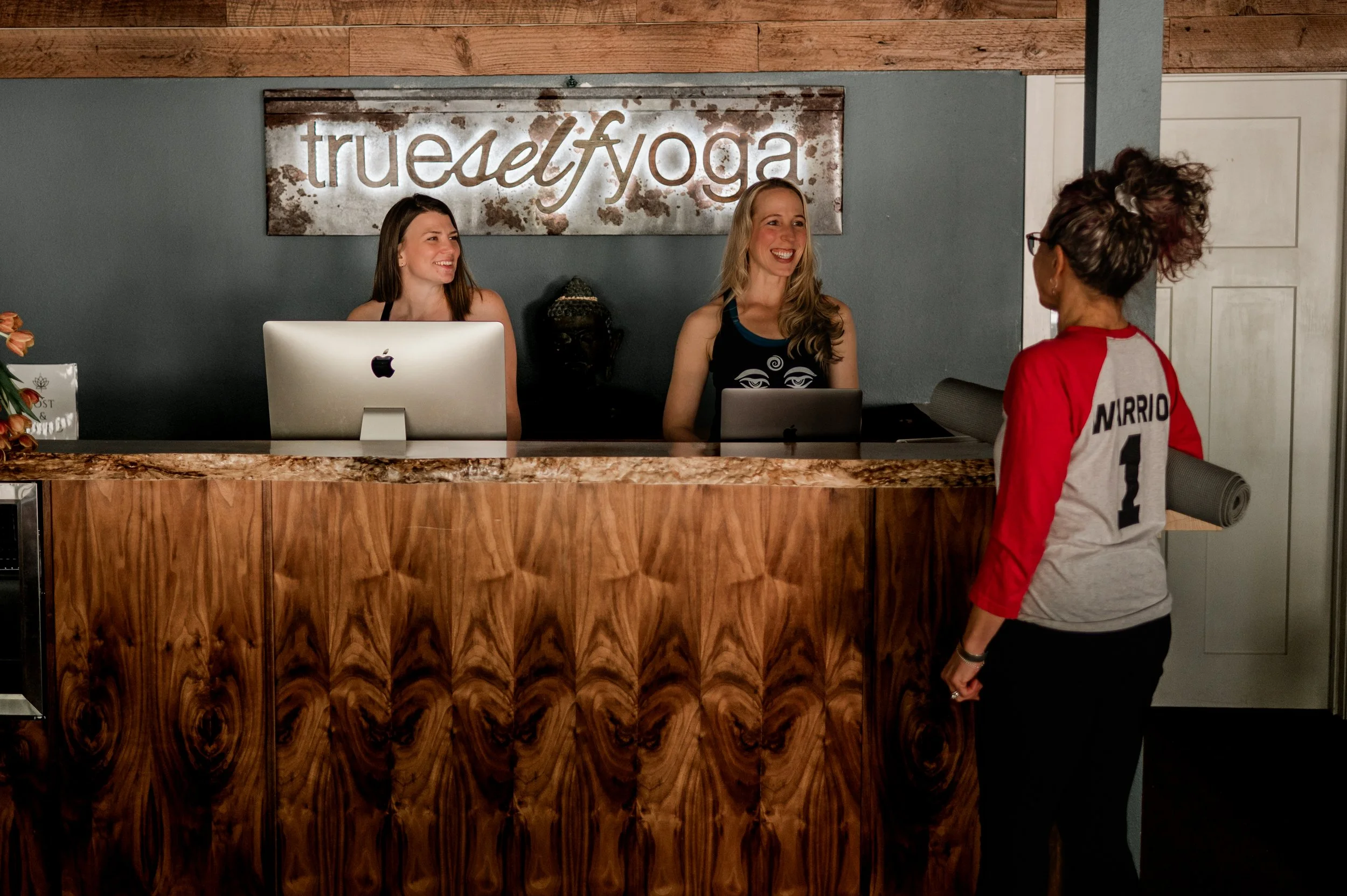 Reception desk at True Self Yoga with two smiling women sitting behind it and a guest standing in front, wearing a red and white sports jersey with the name Mirrio and the number 1 on the back.