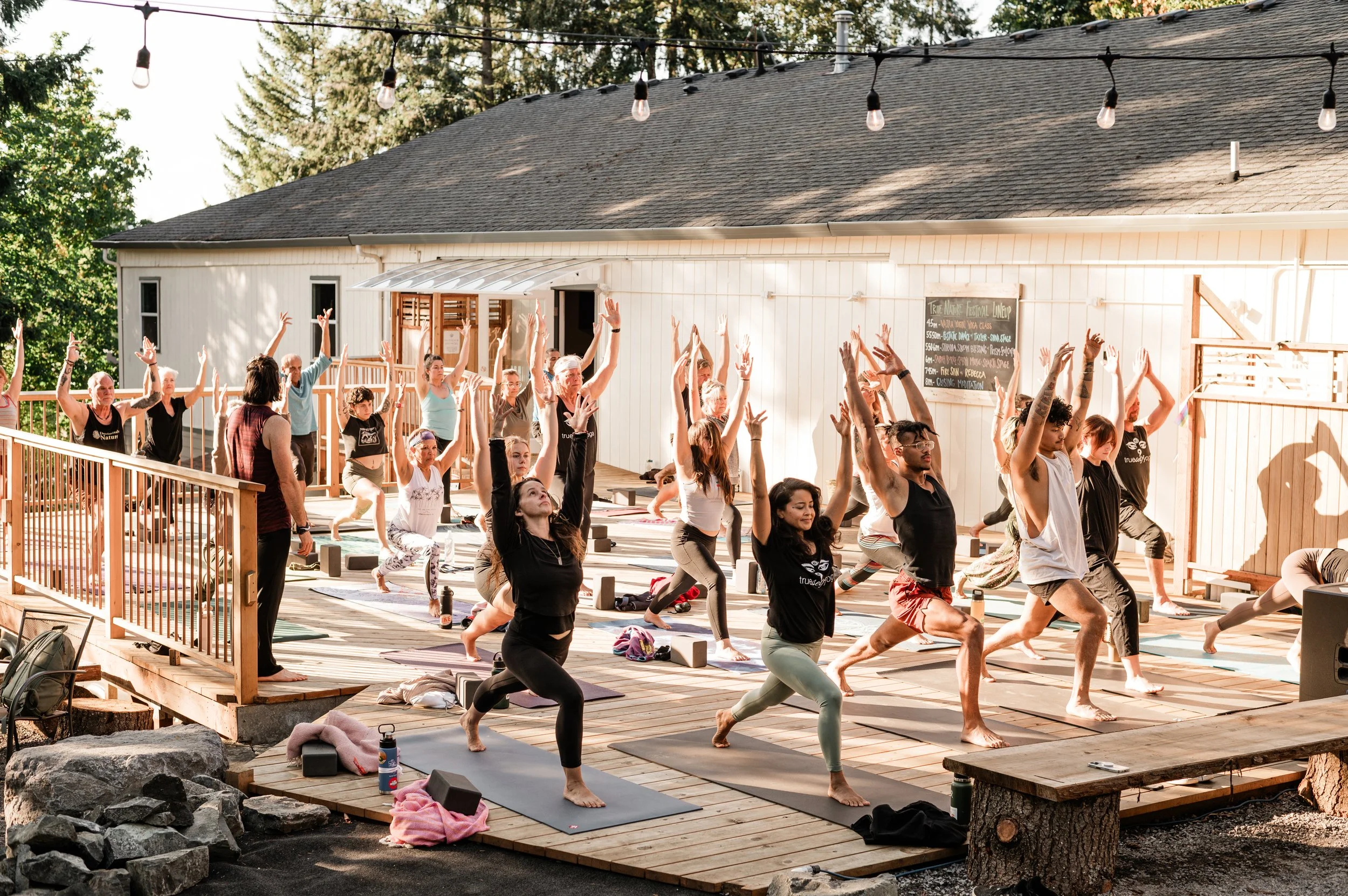 A group of people practicing yoga outdoors on a wooden deck, with some participants in warrior pose and others in downward dog, surrounded by trees and sunset sunlight.